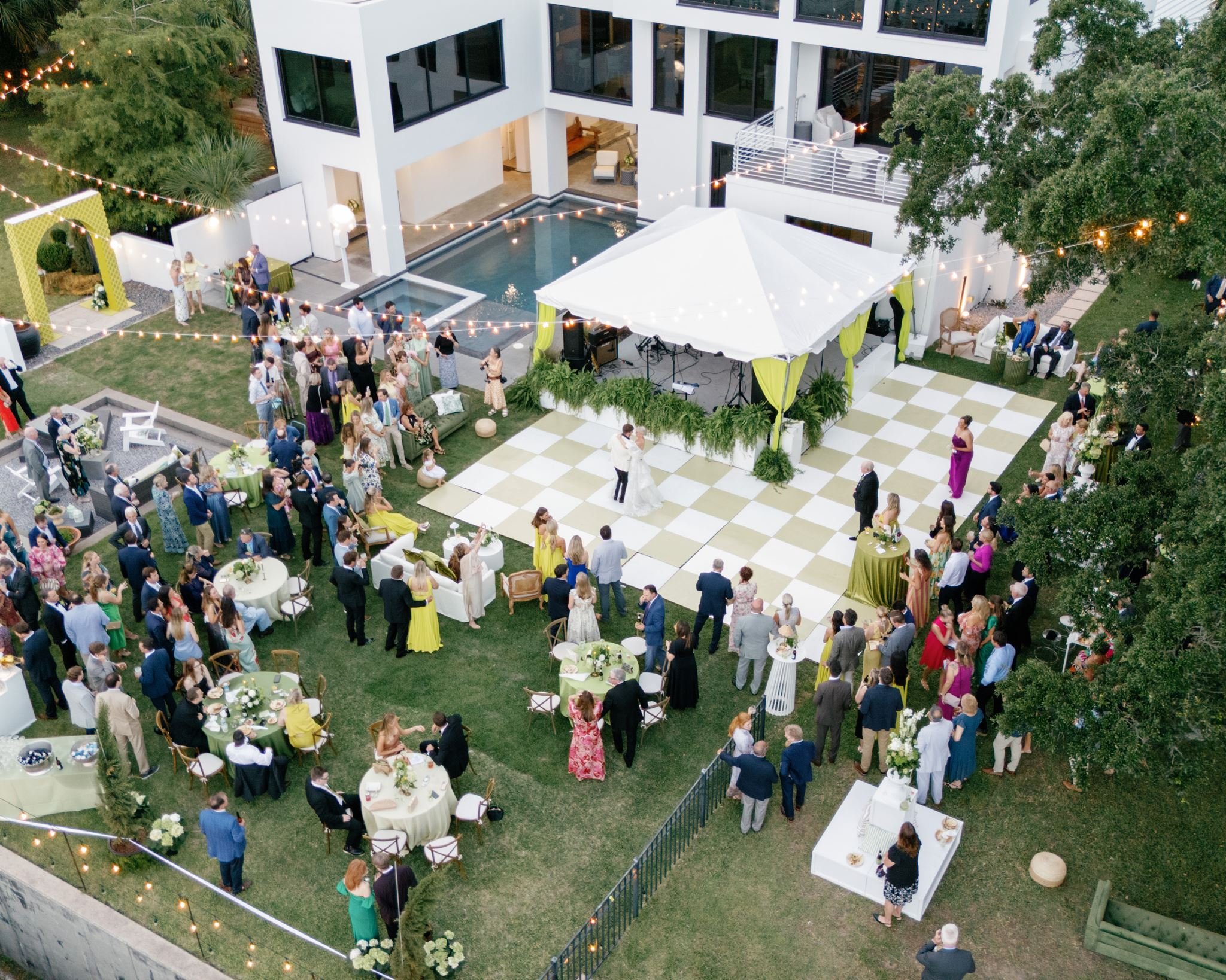 Aerial view of a large outdoor party or wedding reception at a modern white house with a pool, string lights, and various groups of people socializing and dining on tables and chairs.
