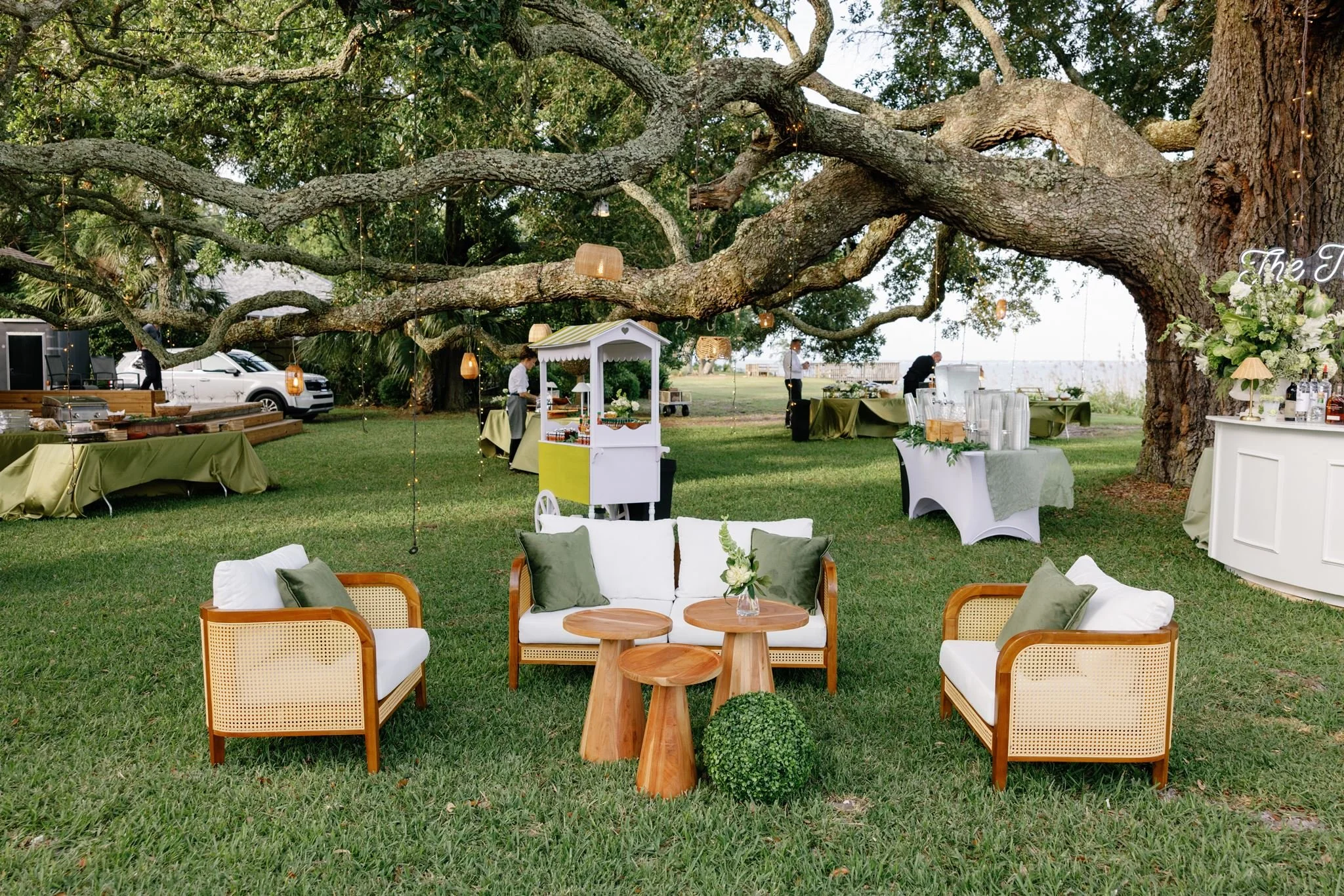 Outdoor wedding reception setup with a large tree, tables covered with green and white linens, and chairs with cushions. String lights hang from the tree branches, and there are decorations and food stations nearby.