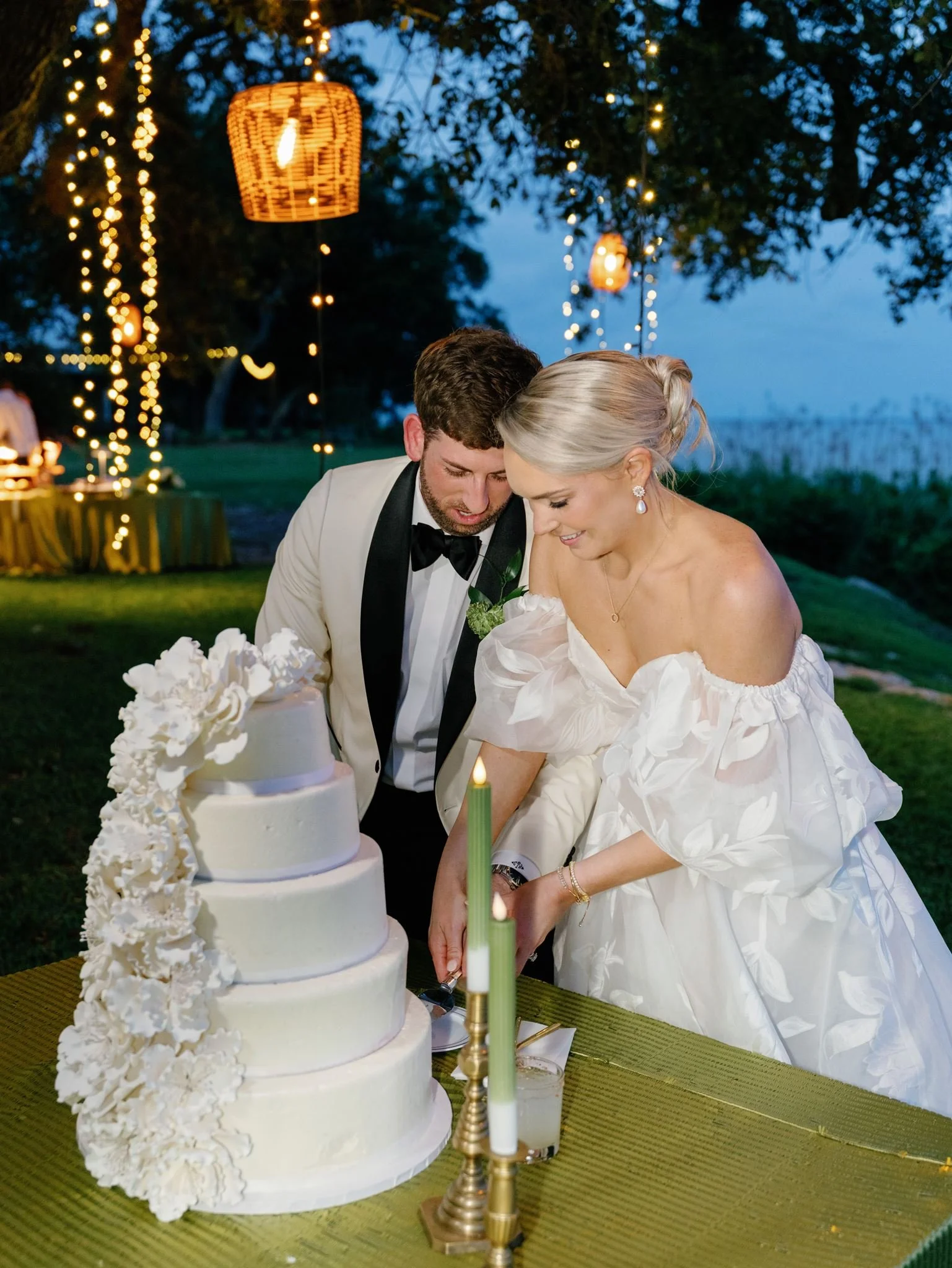 A newlywed couple in white attire cutting a wedding cake outdoors during the evening, decorated with flowers and candles, with hanging lanterns and string lights in the background.