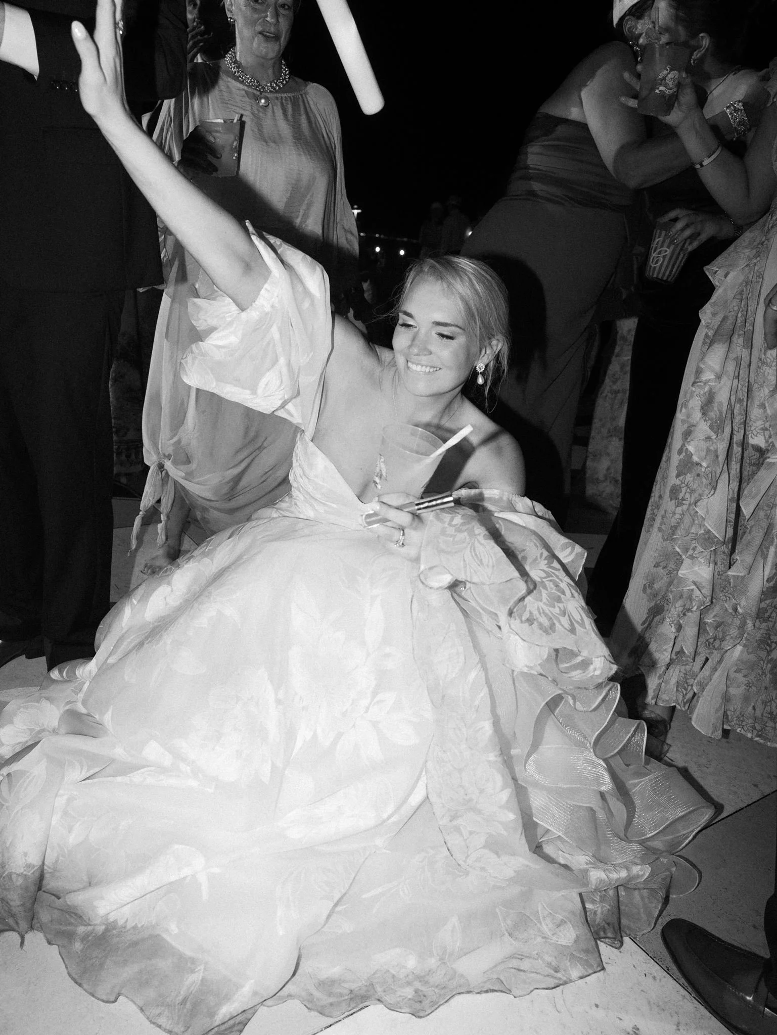 A young woman in a strapless wedding gown smiling while sitting on the floor, surrounded by people at an indoor celebration. She has earrings and a necklace, and a woman standing close by is holding her arm. The scene appears lively with guests holdi