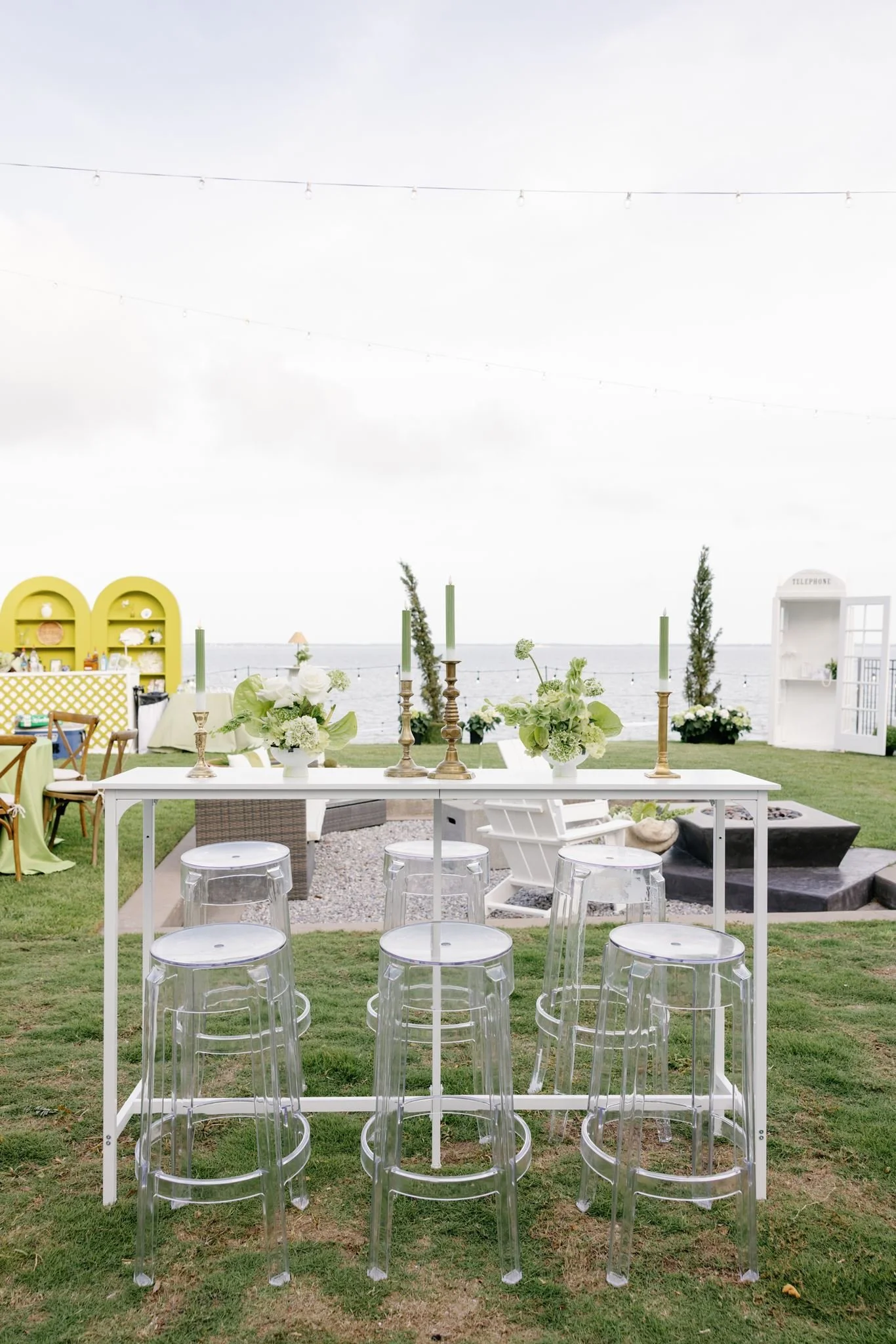 Outdoor event setup with a white table, clear ghost chairs, and floral arrangements, overlooking ocean with string lights hanging overhead.