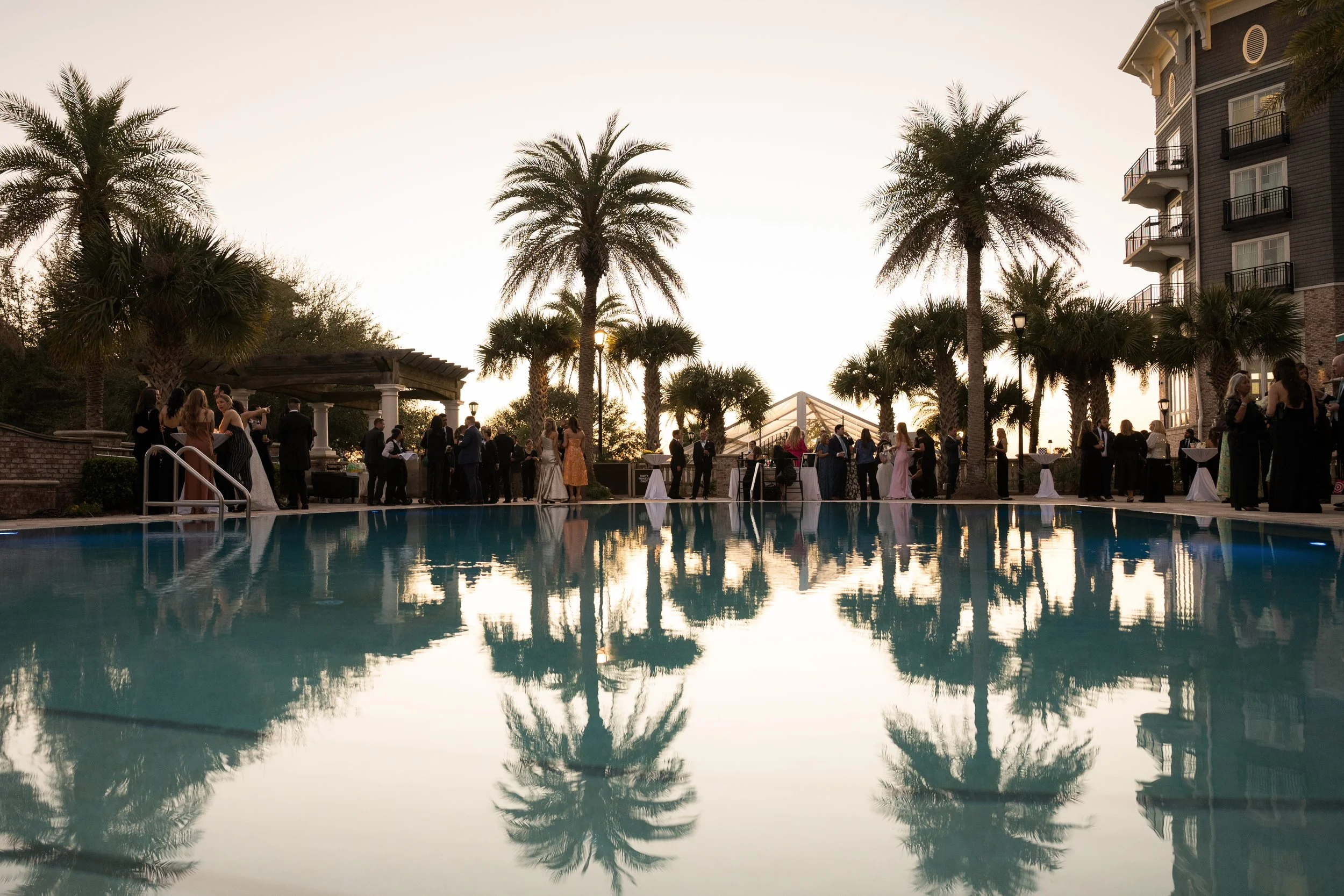 People gathered by a swimming pool at an outdoor event during sunset, with palm trees and a multi-story building in the background.