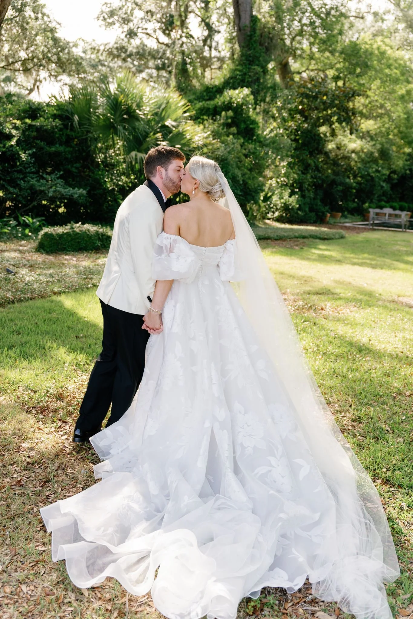 A bride and groom share a kiss outdoors during daytime. The bride is in a white dress with a long train, and the groom is in a white tuxedo jacket with dark trousers. They are holding hands, and the background features trees and green foliage with su
