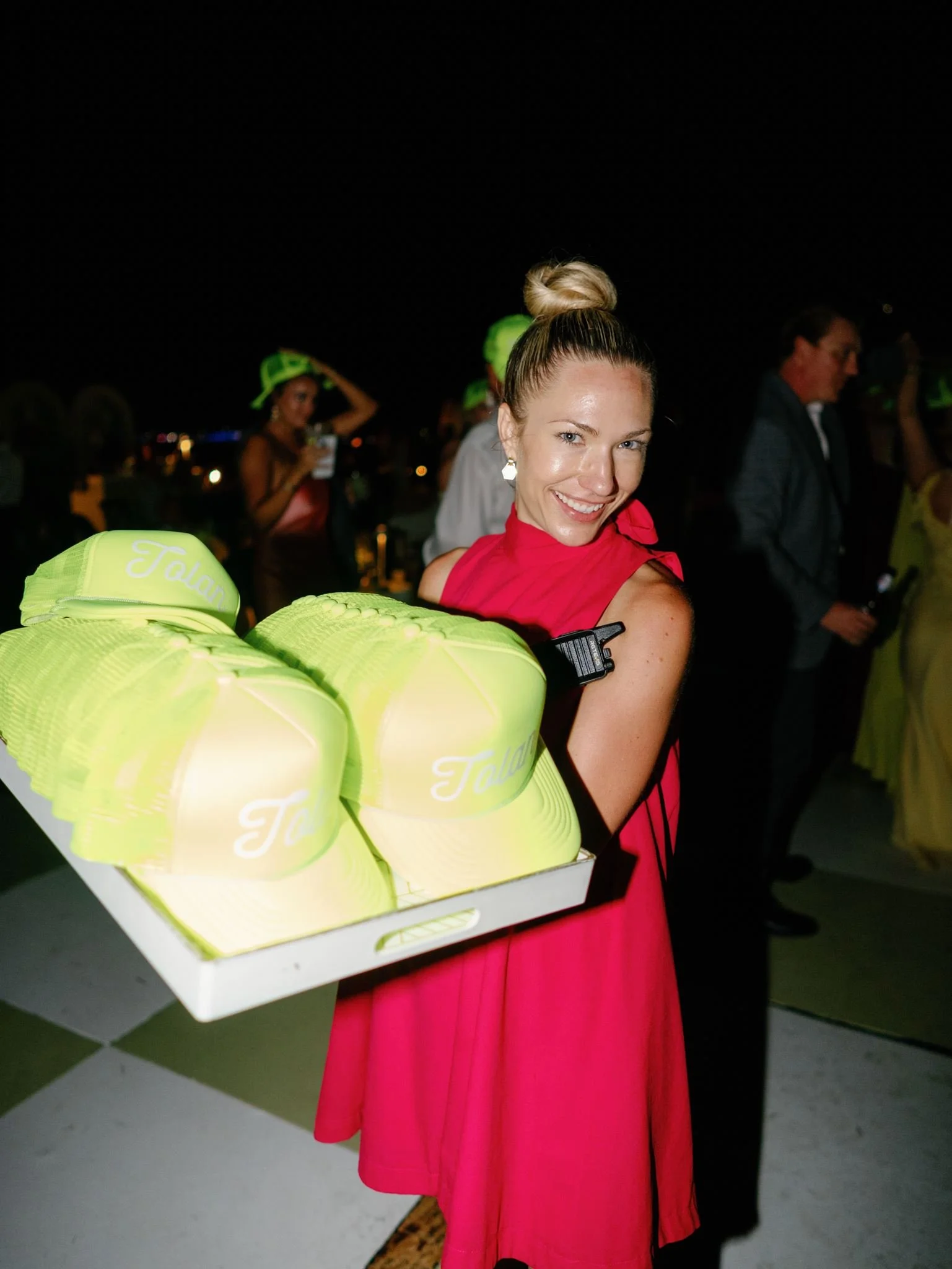 Woman in pink dress holding a tray with neon yellow hats, smiling at the camera at night.