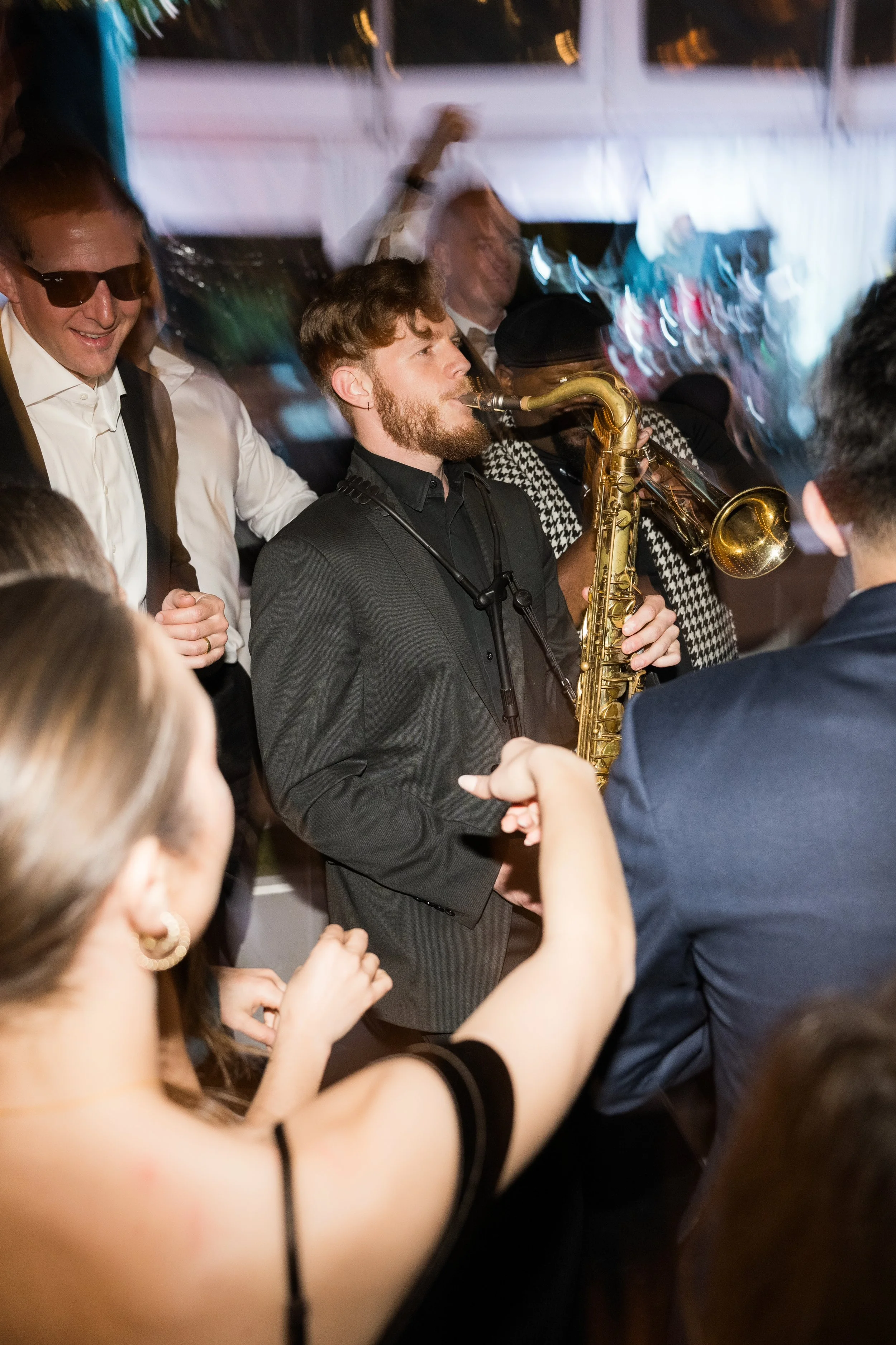 A group of people dancing and playing musical instruments at a lively gathering, including a man playing a saxophone.