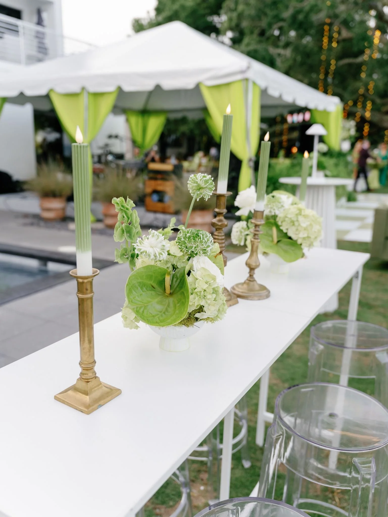 Decorative flower arrangement with green and white blooms, surrounded by three tall green candles on gold candleholders, set on a white table at an outdoor event.