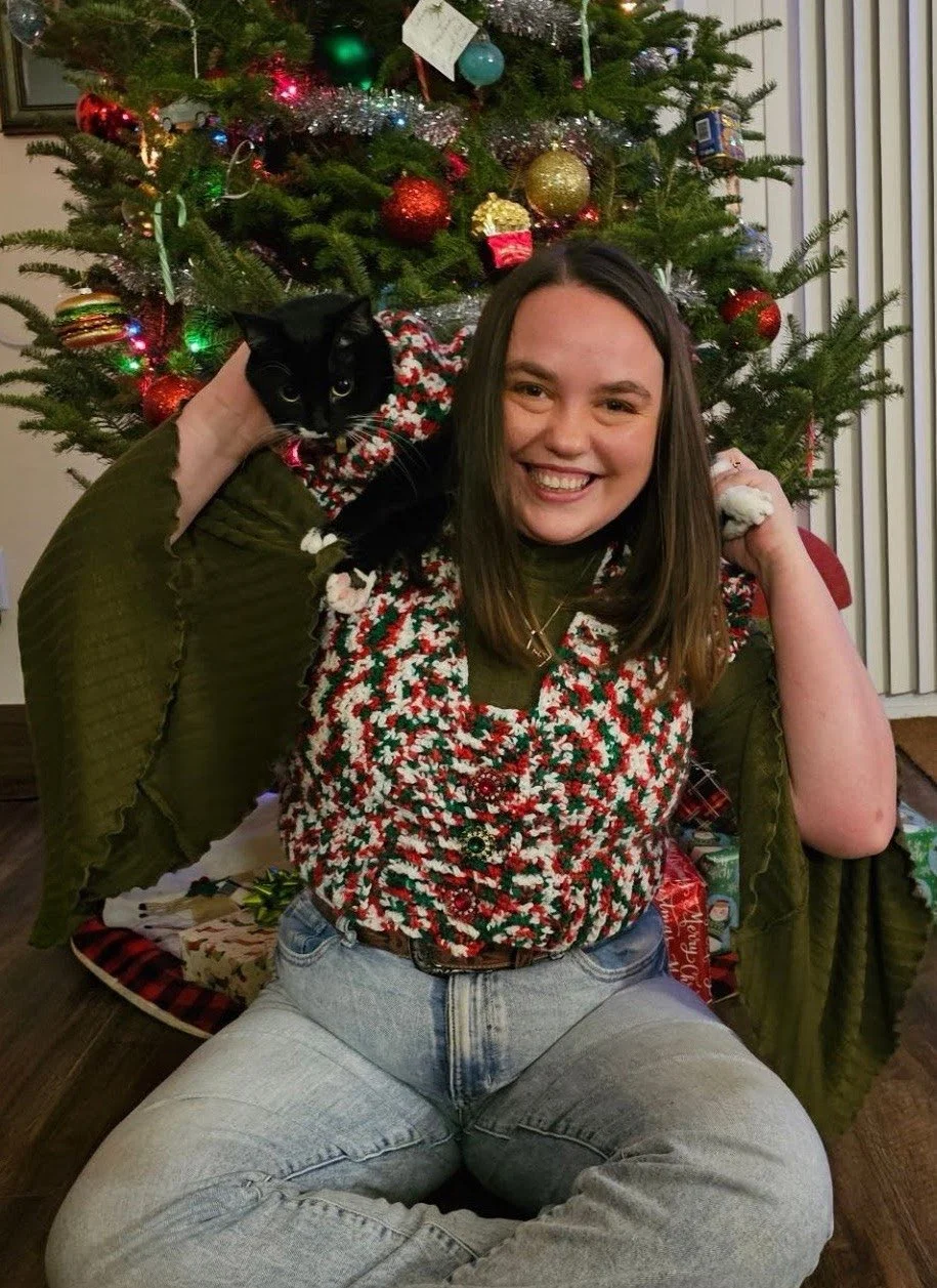 A woman smiling and sitting cross-legged in front of a decorated Christmas tree, holding a black cat on her shoulder. The woman is wearing a festive red, green, and white sweater, and the scene appears to be indoors during Christmas.