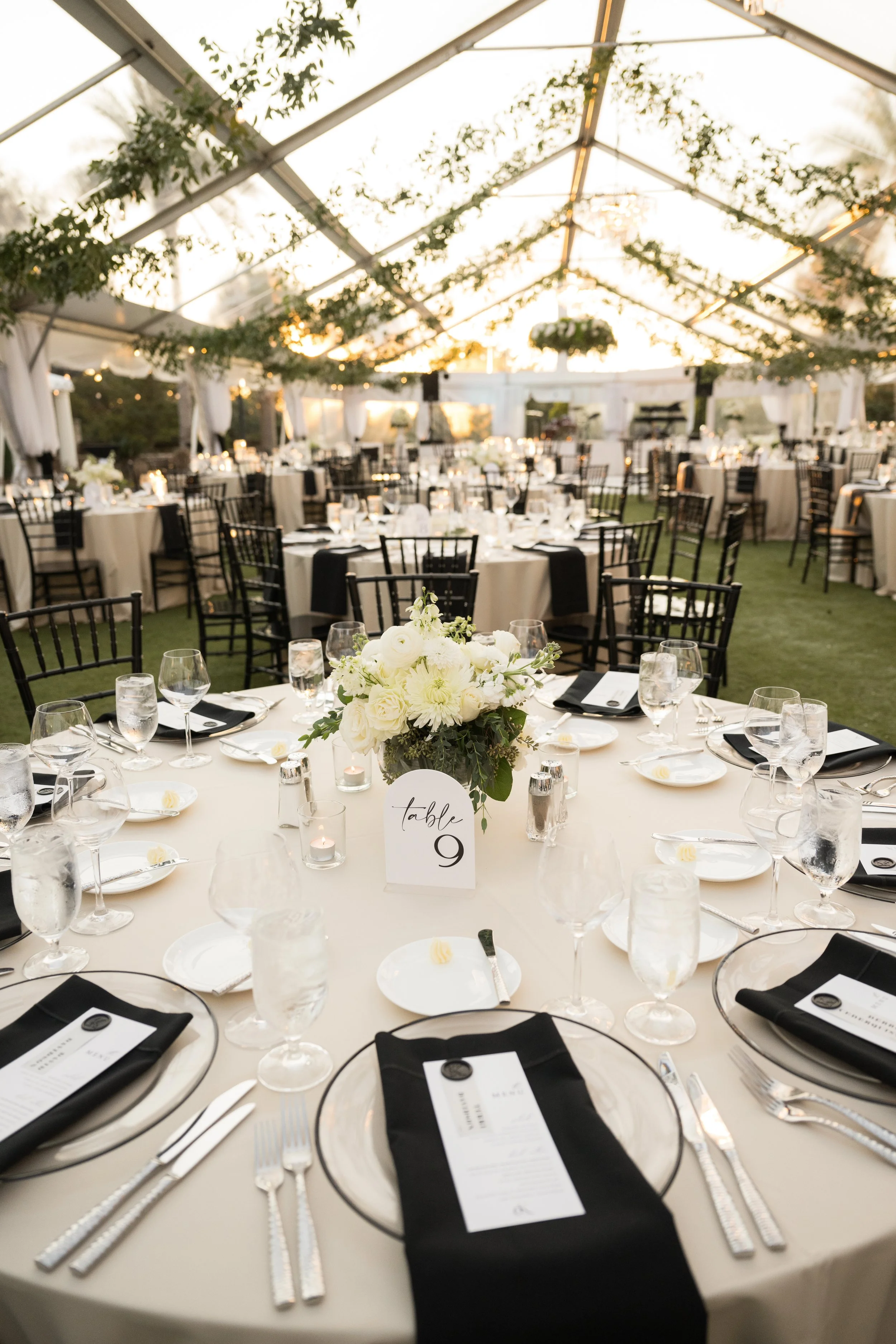 Round banquet table decorated with a white tablecloth, a floral centerpiece of white flowers, surrounded by black napkins, water glasses, and place settings, in an elegant outdoor event space with draped fabric and hanging greenery.