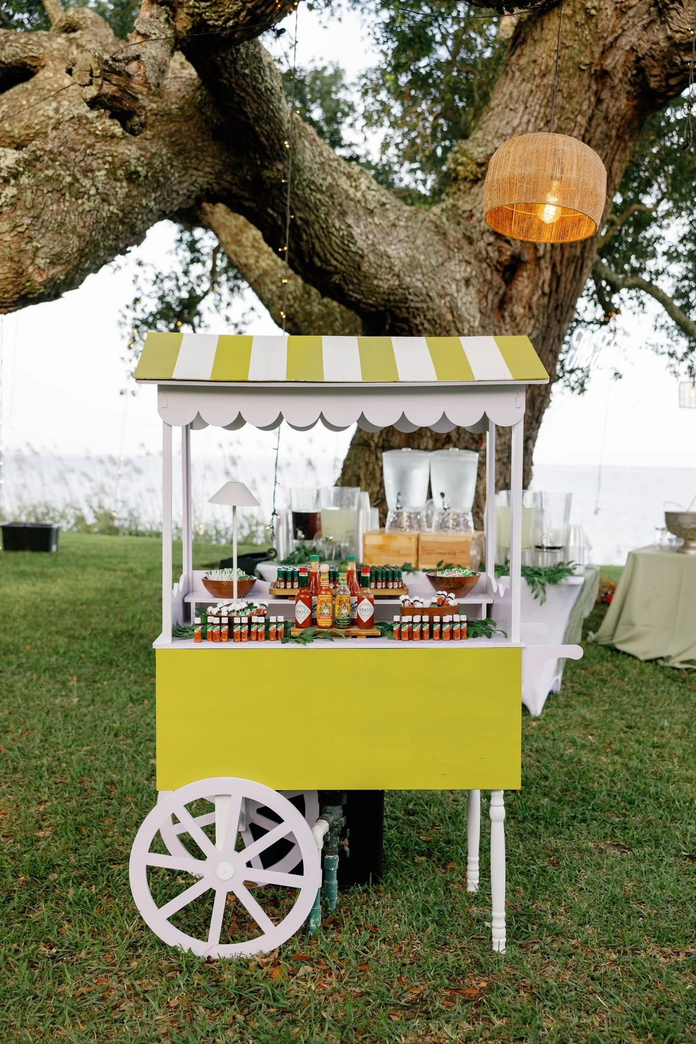 A white and yellow food cart with a scalloped canopy and large white wheels, set outdoors on green grass beneath a large tree, with bottles and condiments displayed on the cart.