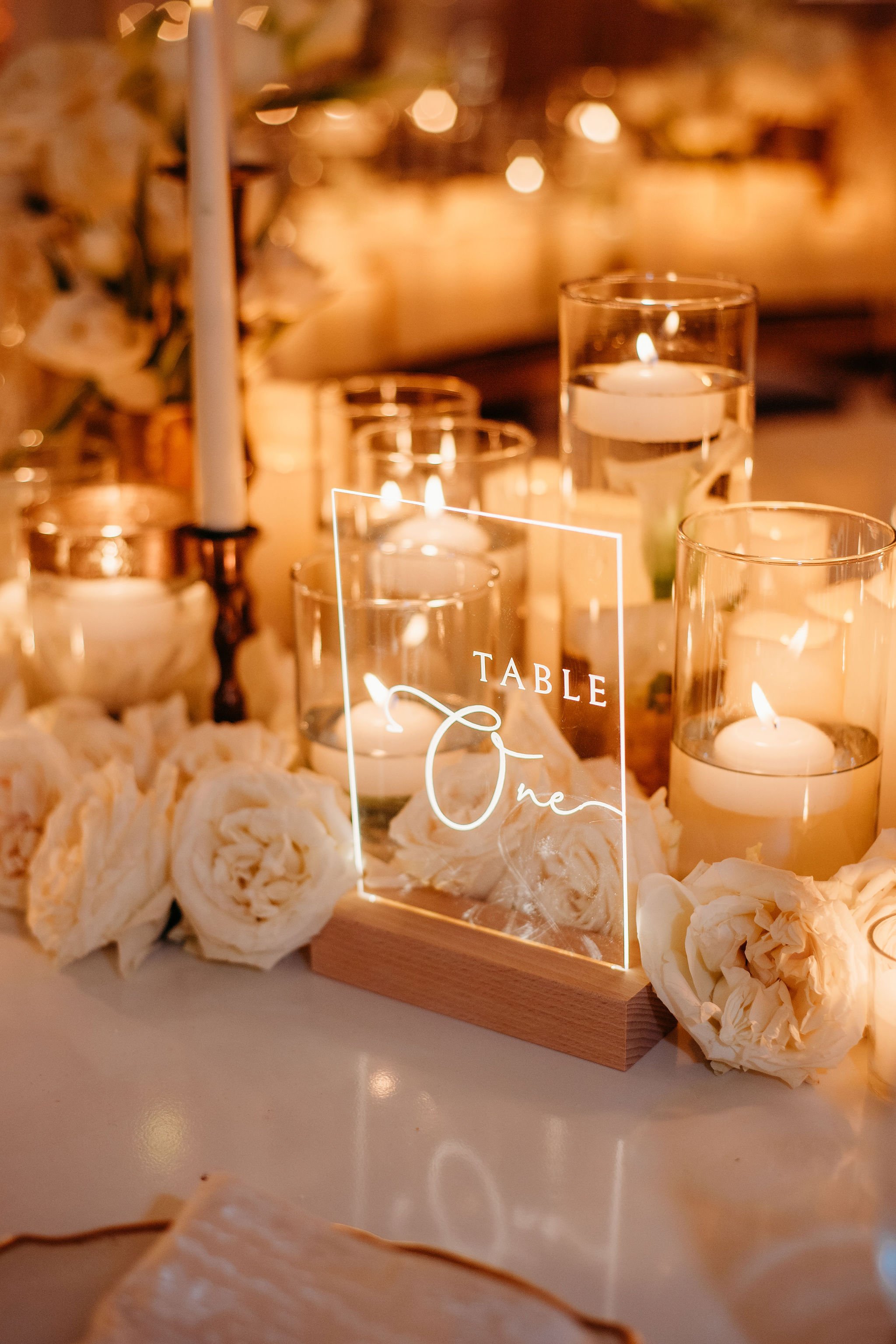 A table centerpiece with white flowers, candles floating in glasses of water, and a decorative sign reading 'Table One' in cursive writing, illuminated by warm candlelight.
