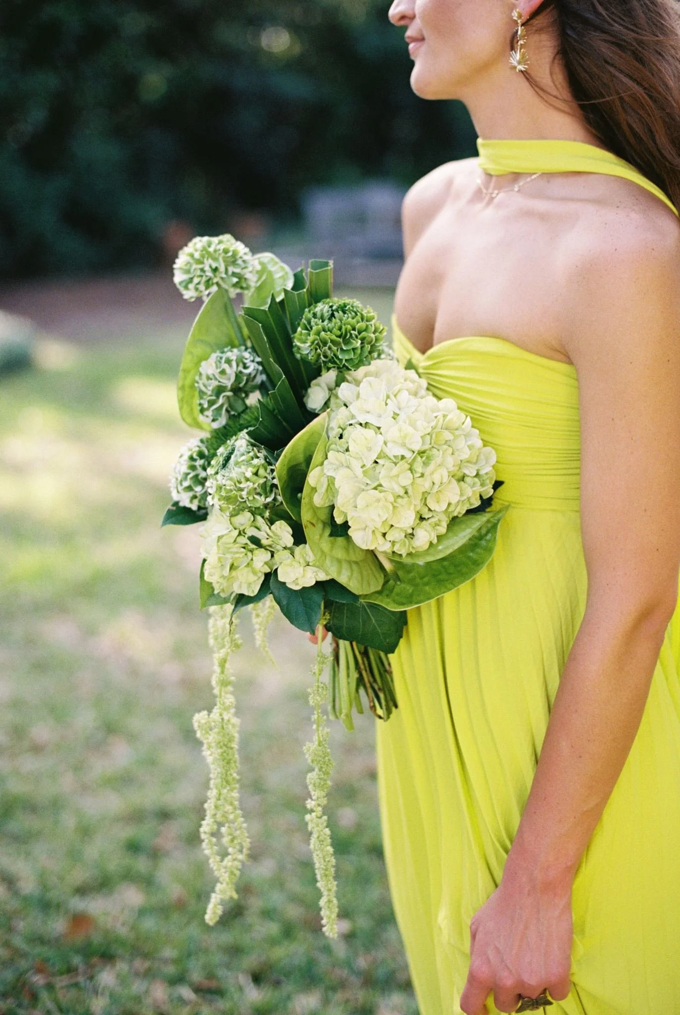 Close-up of a woman in a bright yellow dress holding a bouquet of white and green flowers outdoors. The woman is wearing earrings and a necklace, with sunlight on her face and shoulder. The background is blurred greenery.