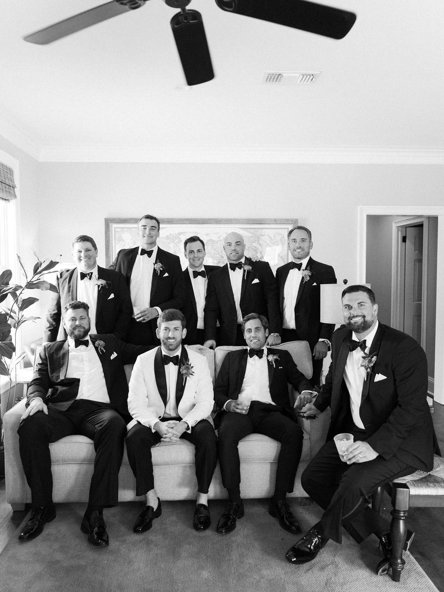 A group of nine men dressed in tuxedos with bow ties, seated on a couch and standing behind it in a living room, celebrating a formal event, possibly a wedding.