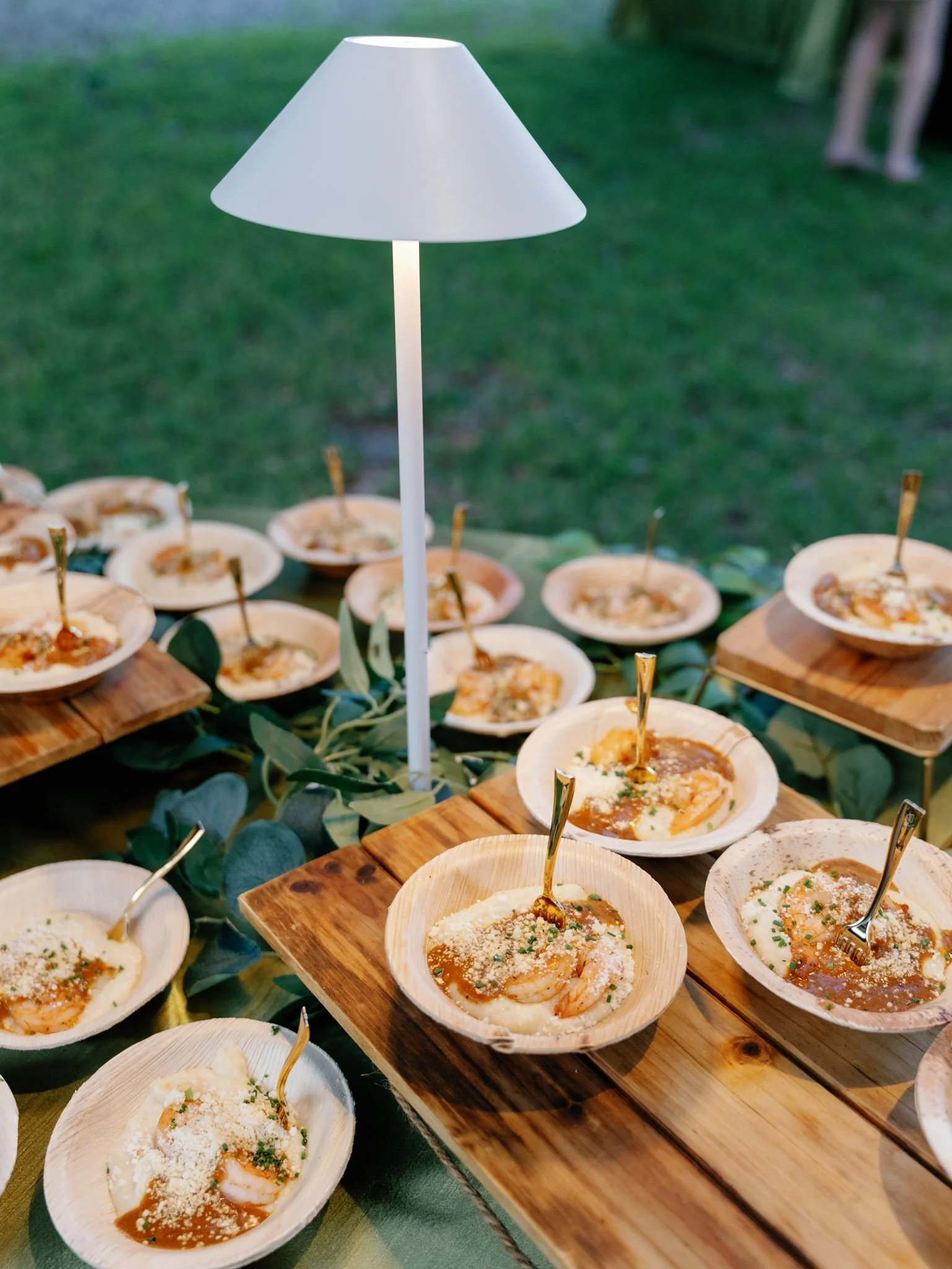 A white lamp with a conical shade standing on a table decorated with wood serving bowls of shrimp and sauce, garnished with cheese and herbs, in an outdoor setting.