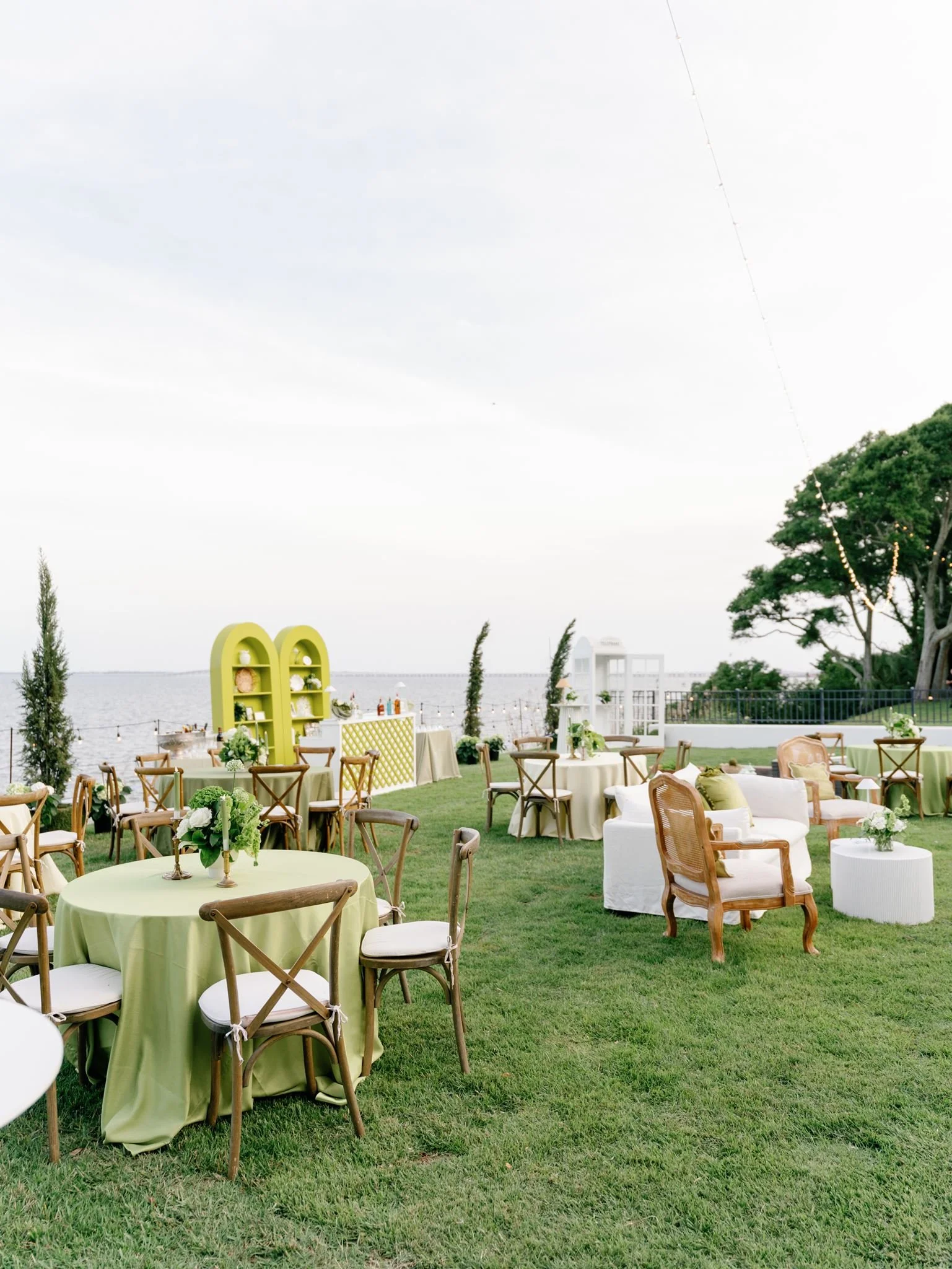 An outdoor event setup by the water featuring round tables with green tablecloths, wooden chairs, floral centerpieces, and a yellow decorative shelf with plants and accessories, under a cloudy sky with string lights and trees.