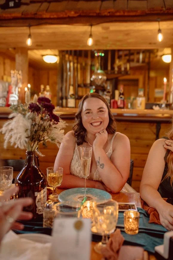 A woman with shoulder-length brown hair, wearing a sleeveless beige dress, smiling and sitting at a dinner table with a champagne glass in front of her. The table has a vase with flowers, candles, and plates. The setting is a rustic wooden restaurant or bar.