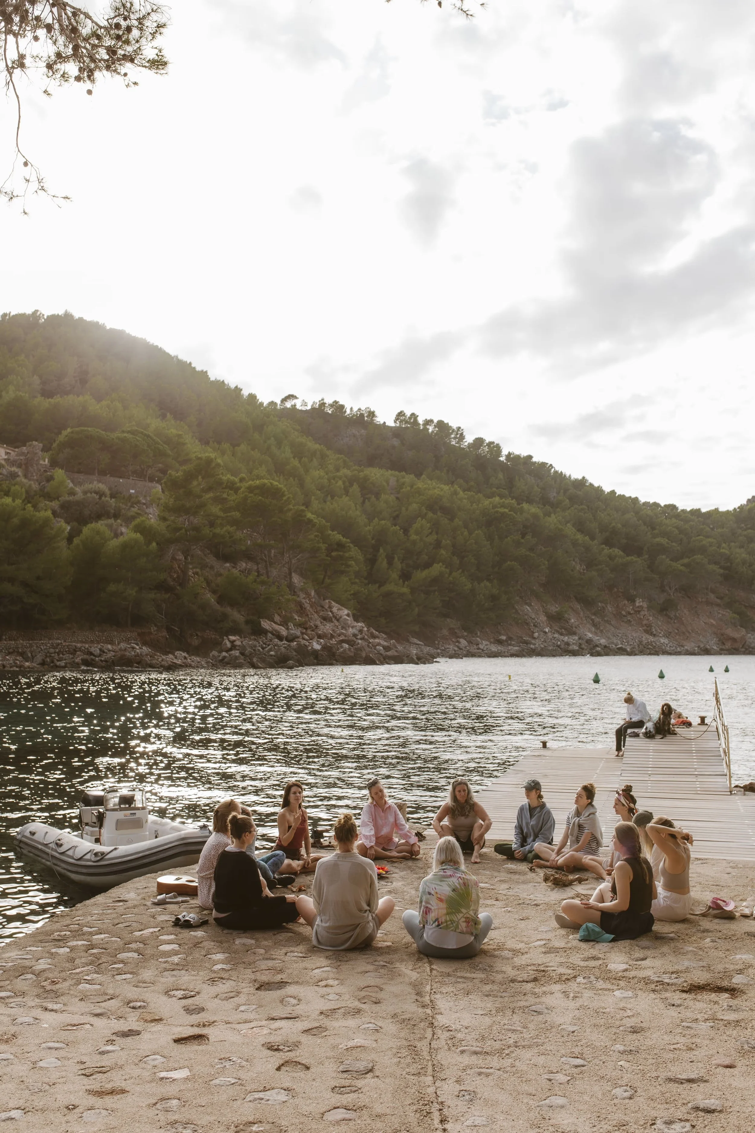 A group of people sitting in a circle on a paved area by a body of water, with a dock extending into the water. The scene is surrounded by lush green hills and trees, with some boats visible in the water.