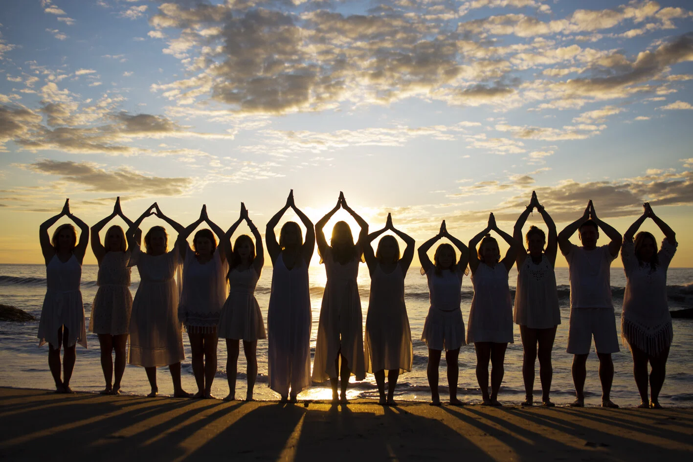 A group of people standing on the beach at sunset, practicing yoga with their hands in prayer position above their heads, silhouetted against the sky and ocean.