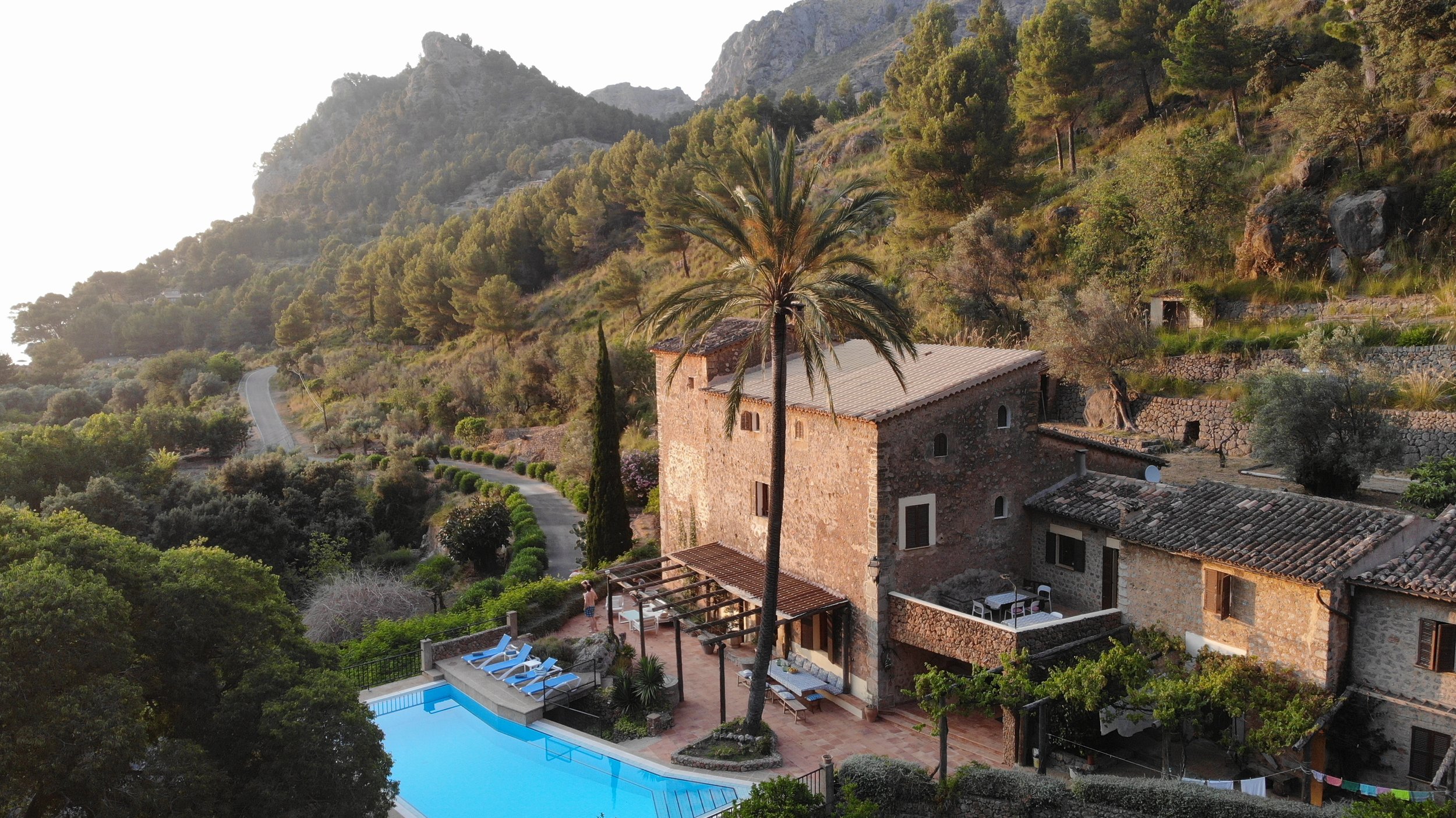 A hillside stone house with a red-tiled roof, surrounded by lush greenery including a tall palm tree, near a swimming pool with lounge chairs, set in a mountainous landscape with trees and winding road.