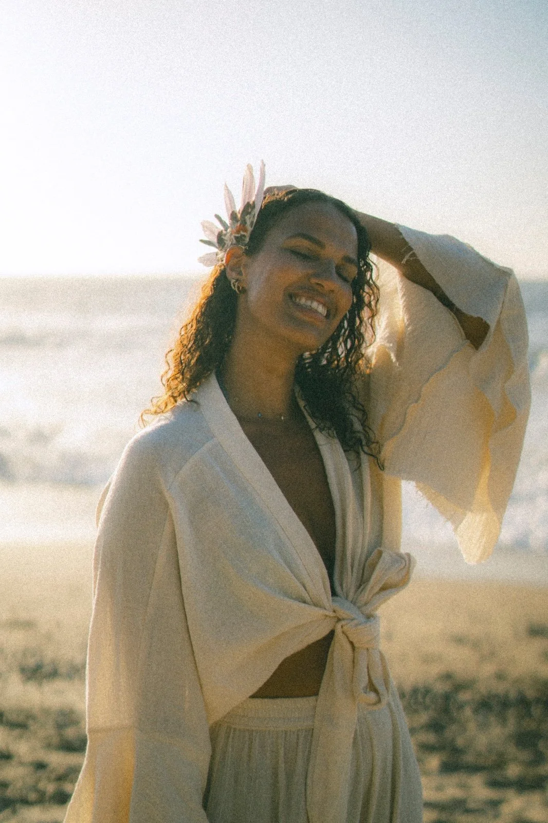 A woman on the beach, squinting and smiling, wearing a light-colored, tied-up cover-up dress, with a floral headband and earrings, with ocean waves in the background.