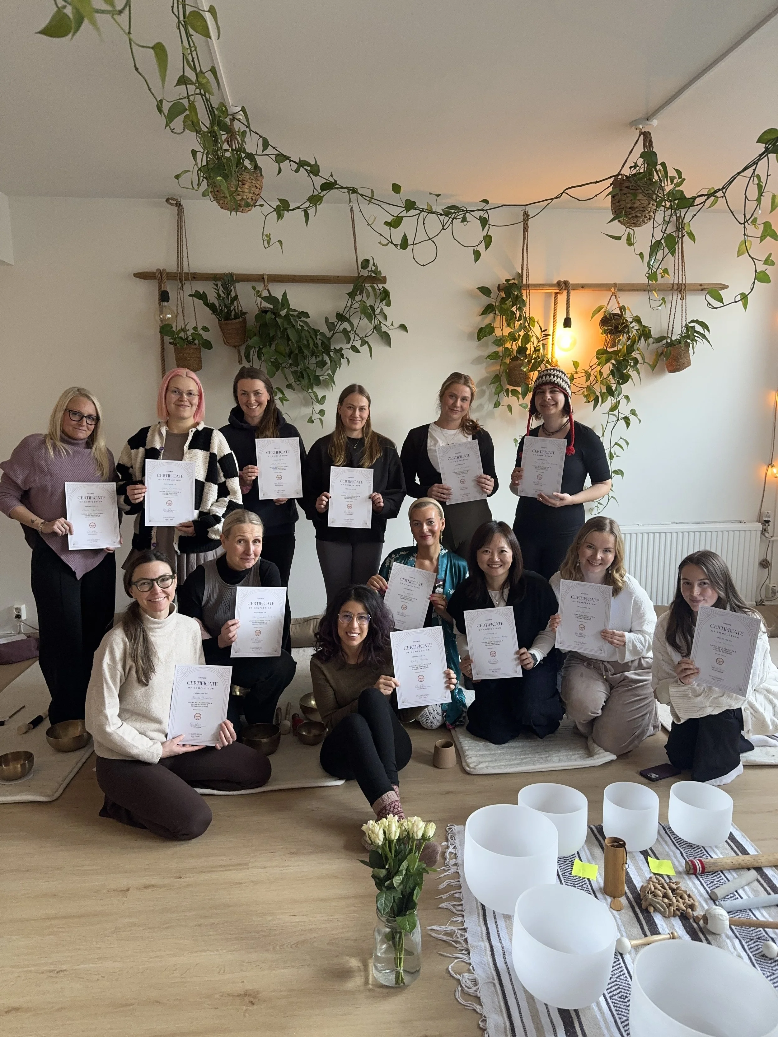 Group of women holding certificates in a decorated indoor space with hanging plants, soft lighting, and singing bowls on the floor.