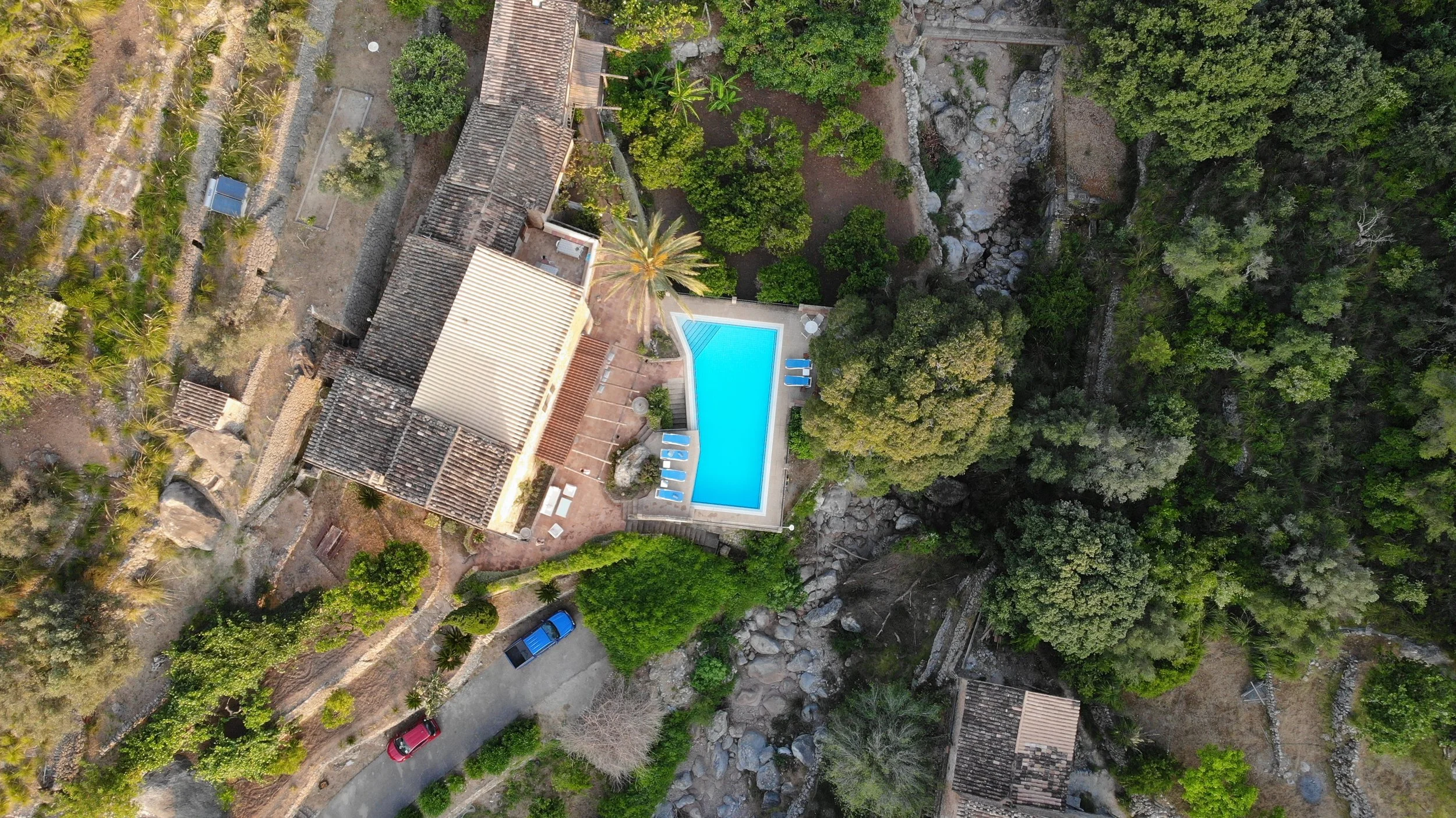 An aerial view of a property with a swimming pool, surrounding greenery, trees, and a few vehicles parked on a driveway.