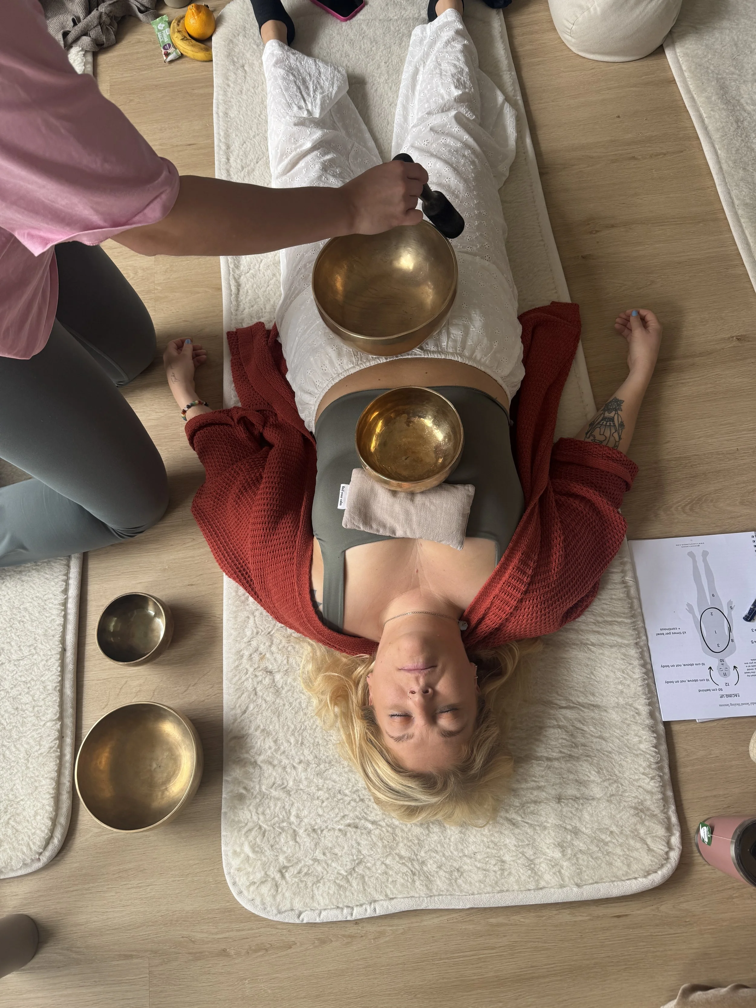 A woman lying on her back on a mat with her eyes closed, with bowls placed on her chest and stomach, during a sound therapy or relaxation session. A person is holding a singing bowl near her abdomen. There are additional bowls on the floor nearby, al