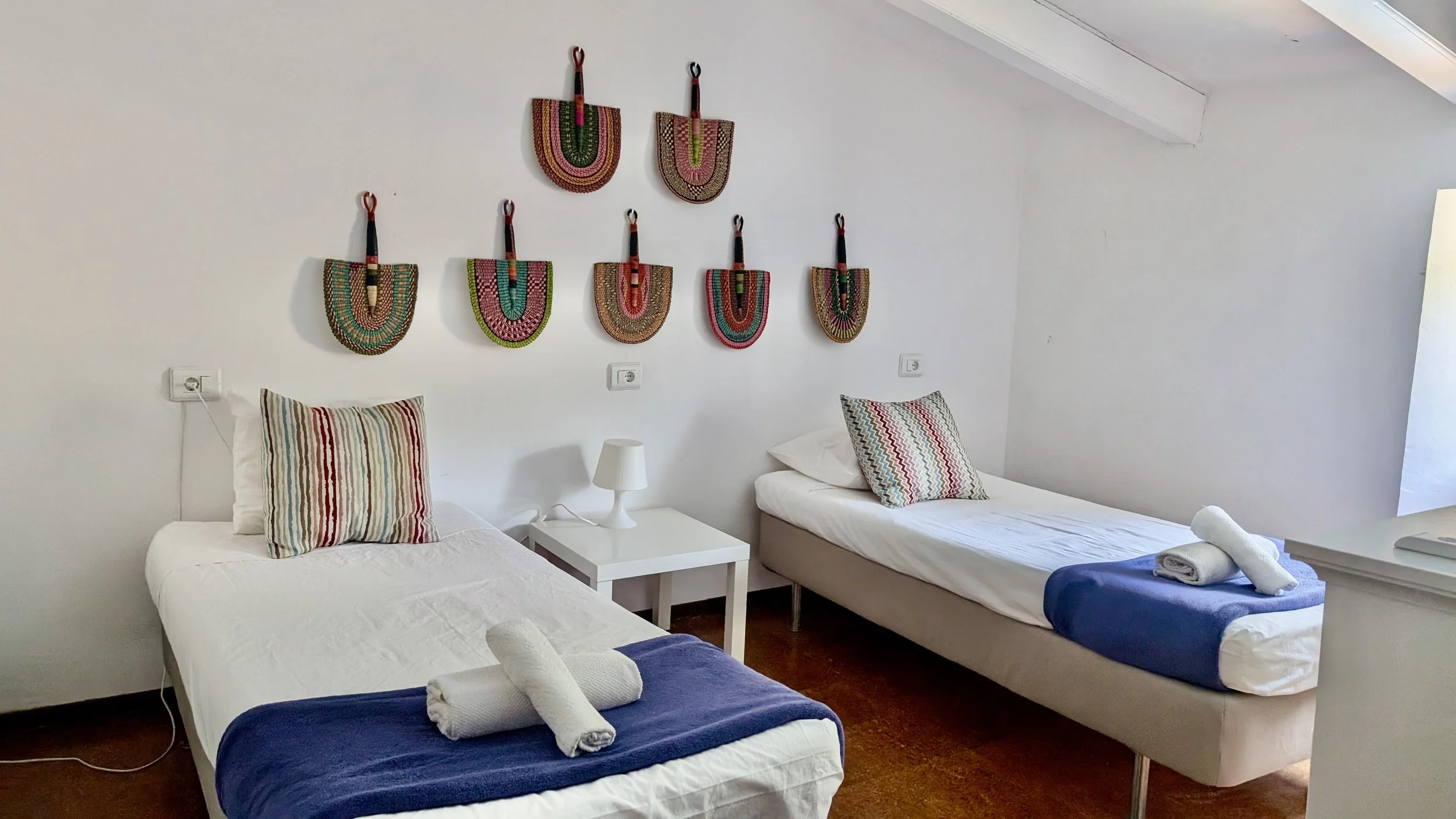 A room with two single beds each with a pillow and rolled towels at the foot, a small white nightstand with a lamp between the beds, and colorful woven wall hangings above the beds.