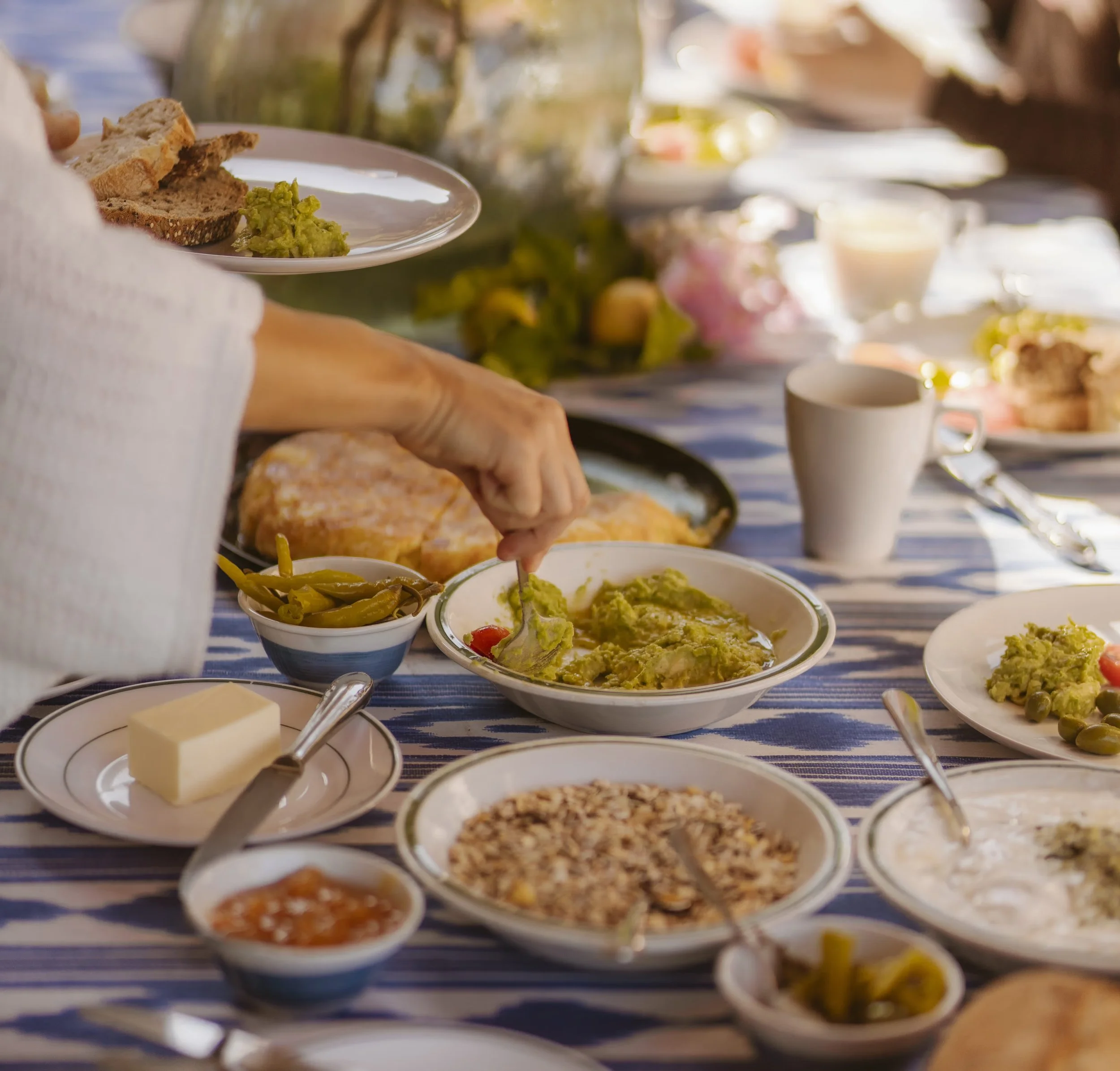 A person serving guacamole at a breakfast or brunch table with various dishes including bread, green beans, cheese, and cereals, set on a striped tablecloth outdoors.