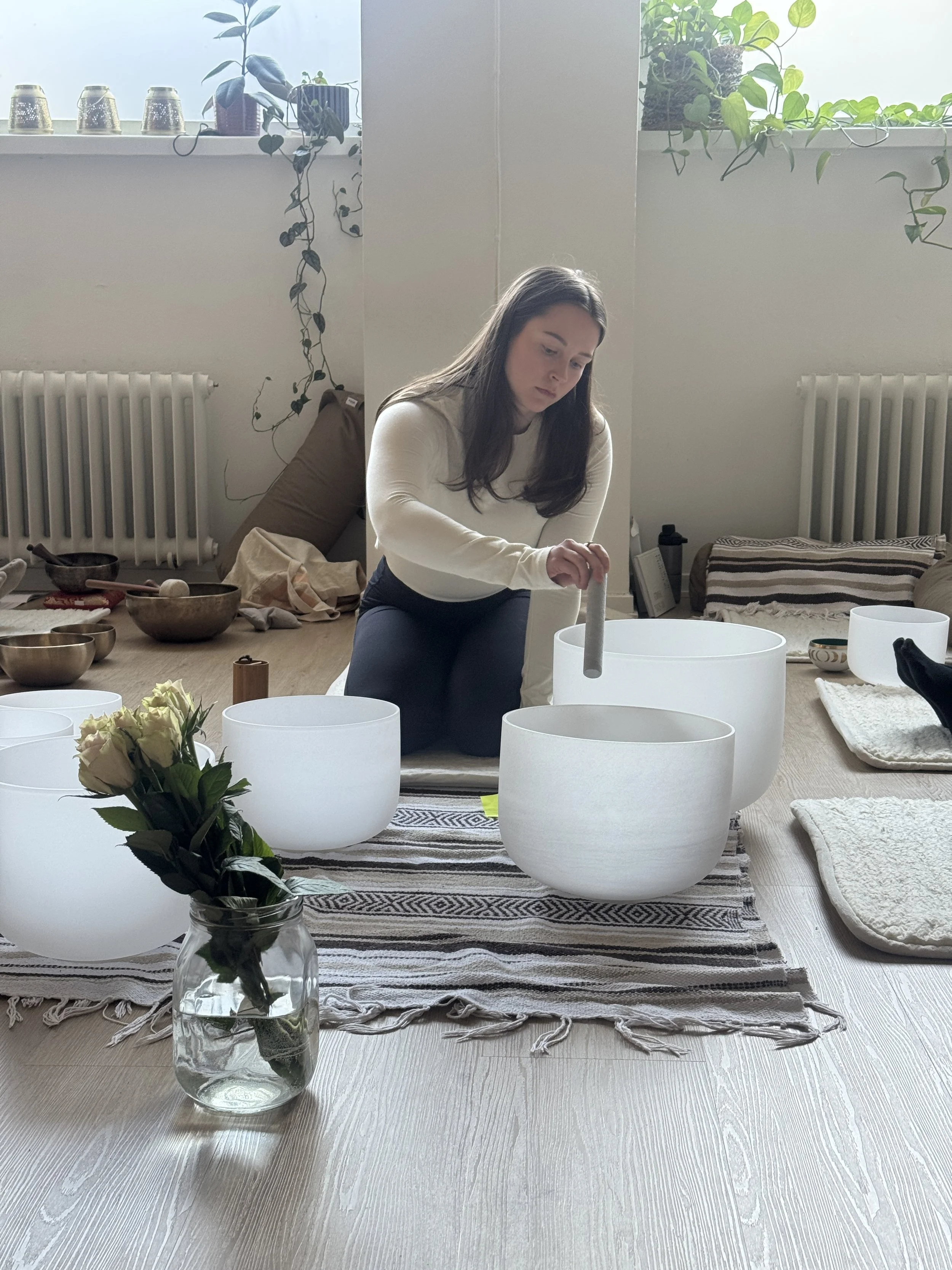A woman is sitting on the floor playing crystal singing bowls on a table with a striped cloth, with a window behind her adorned with potted plants and decorative cups, creating a peaceful, cozy atmosphere.