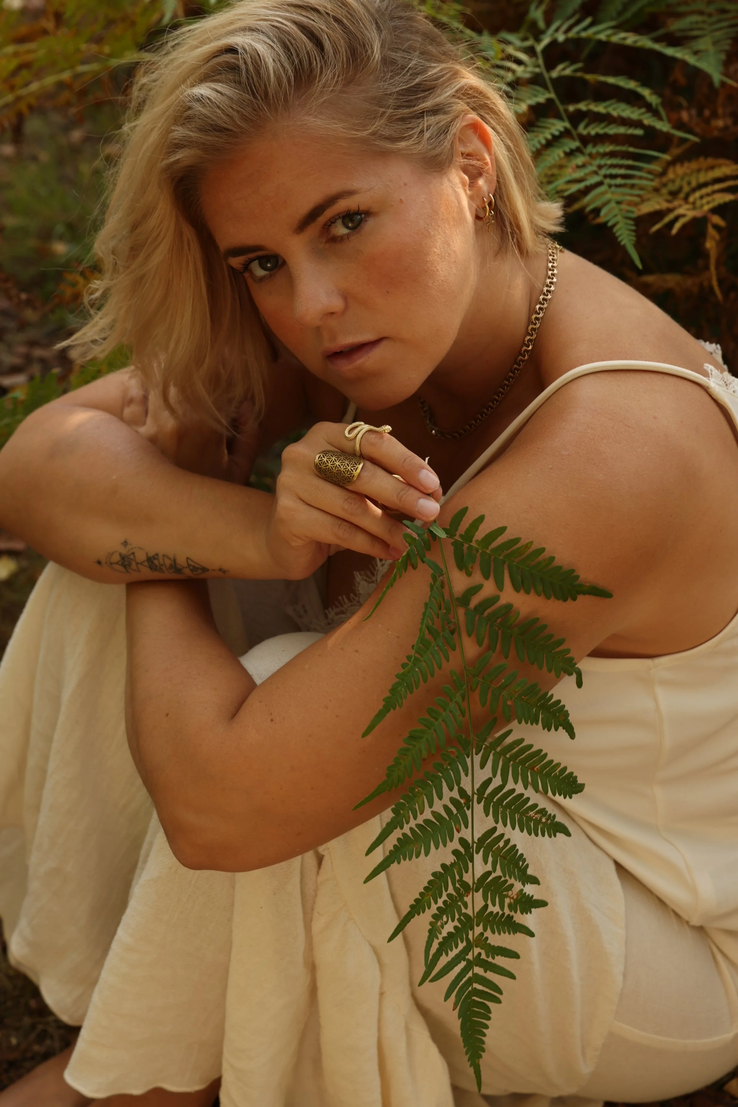 A woman with blonde hair, wearing jewelry, sitting outdoors amidst ferns and natural foliage, looking directly at the camera.