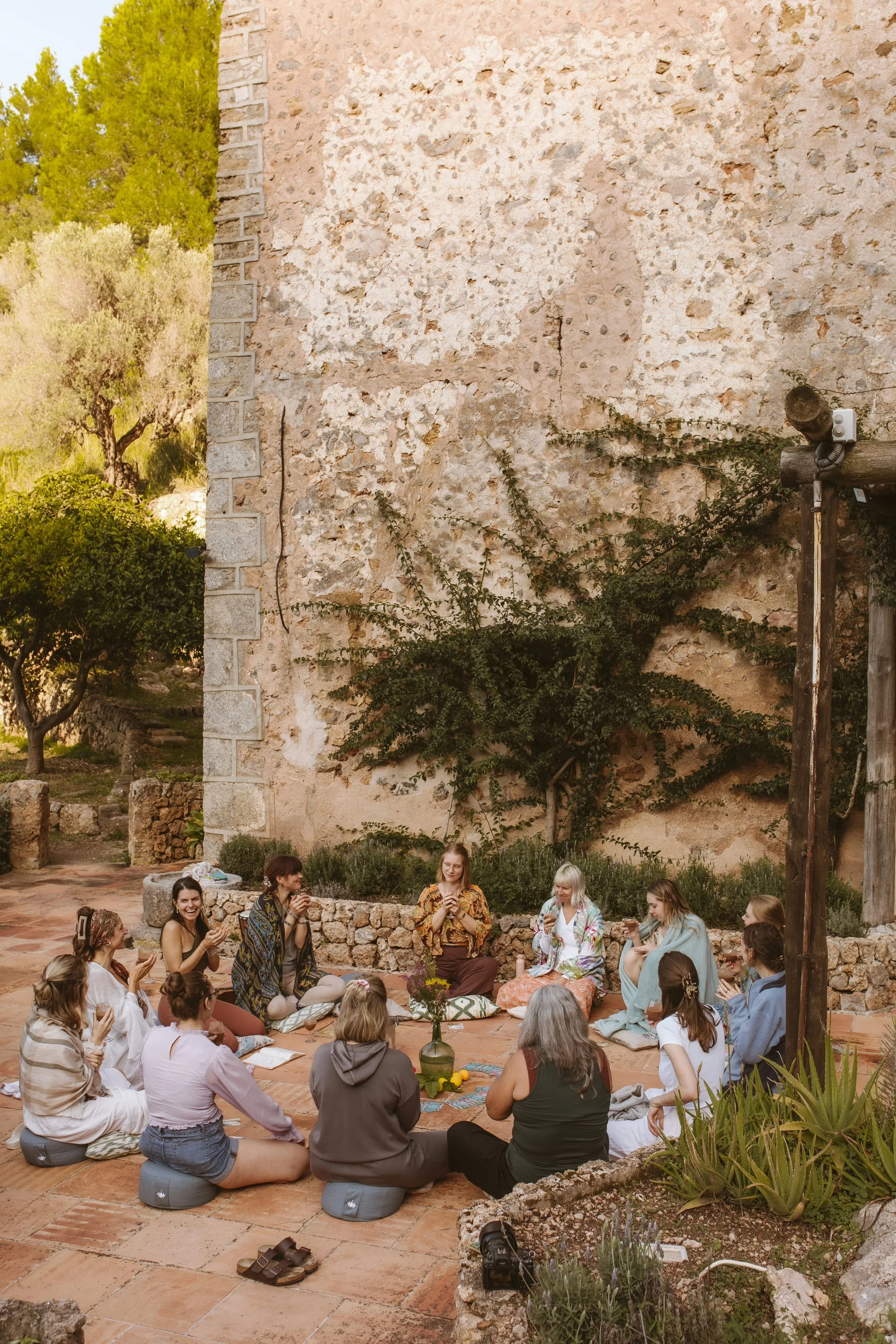 Group of people sitting in a circle outdoors, engaging in a spiritual or meditation session, with plants and rustic wall background.