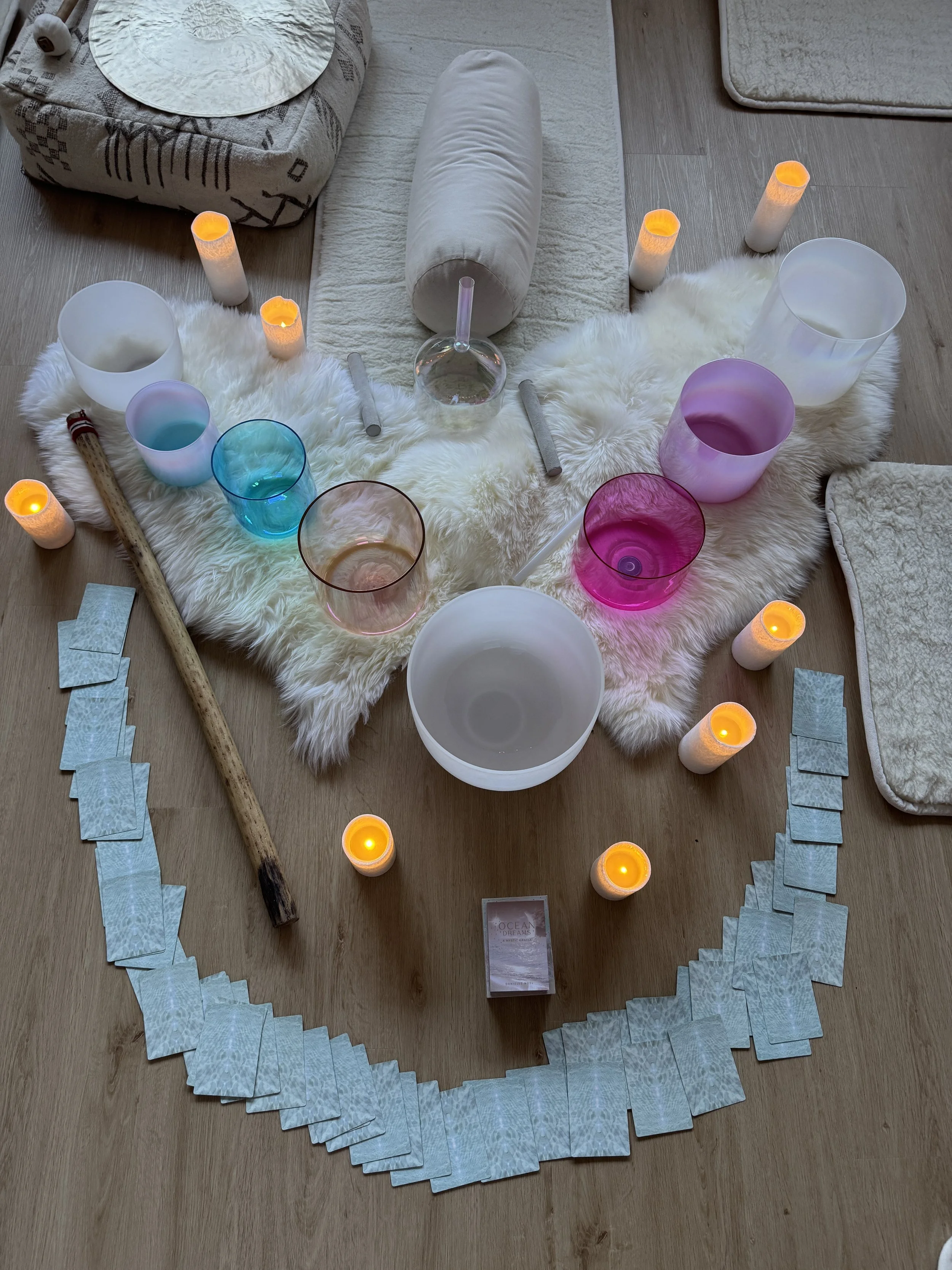 A cozy setup with a white furry rug, several lit candles, colorful singing bowls, a large white bowl, cards, a wooden stick, and a glass instrument arranged in a semi-circle, suggesting a meditation or relaxation session.