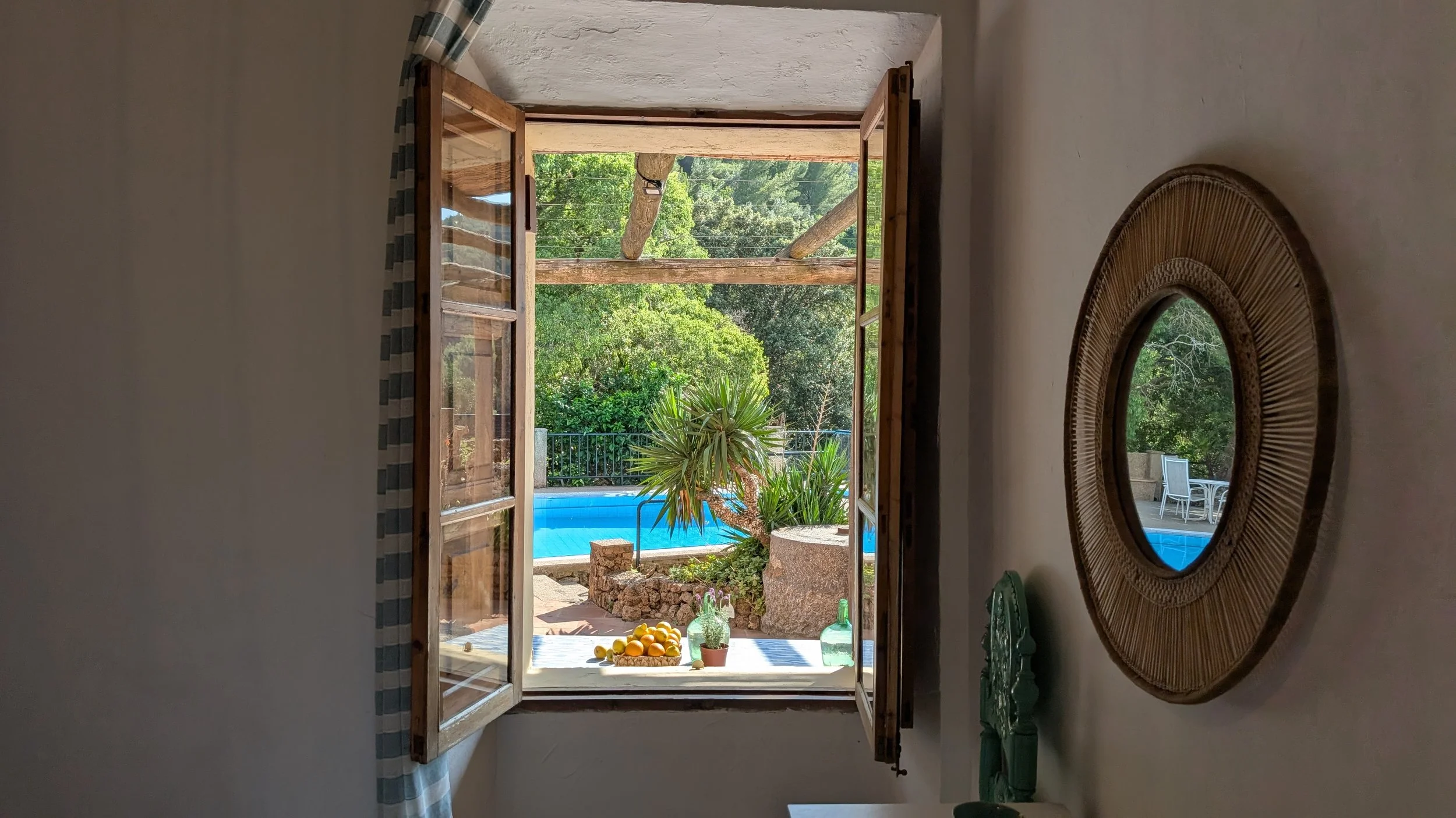 View through an open window showing a backyard with a swimming pool, tropical plants, a wooden pergola, and outdoor furniture.