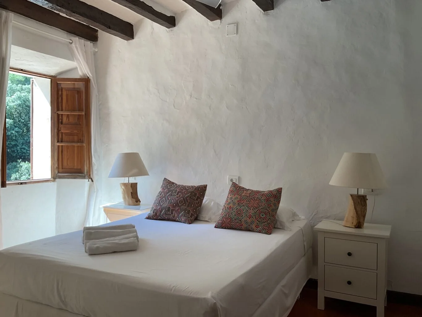 A cozy bedroom with a white bed, decorative pillows, and a white nightstand with a lamp, open window with wooden shutters, white curtains, textured white walls, and exposed wooden ceiling beams.