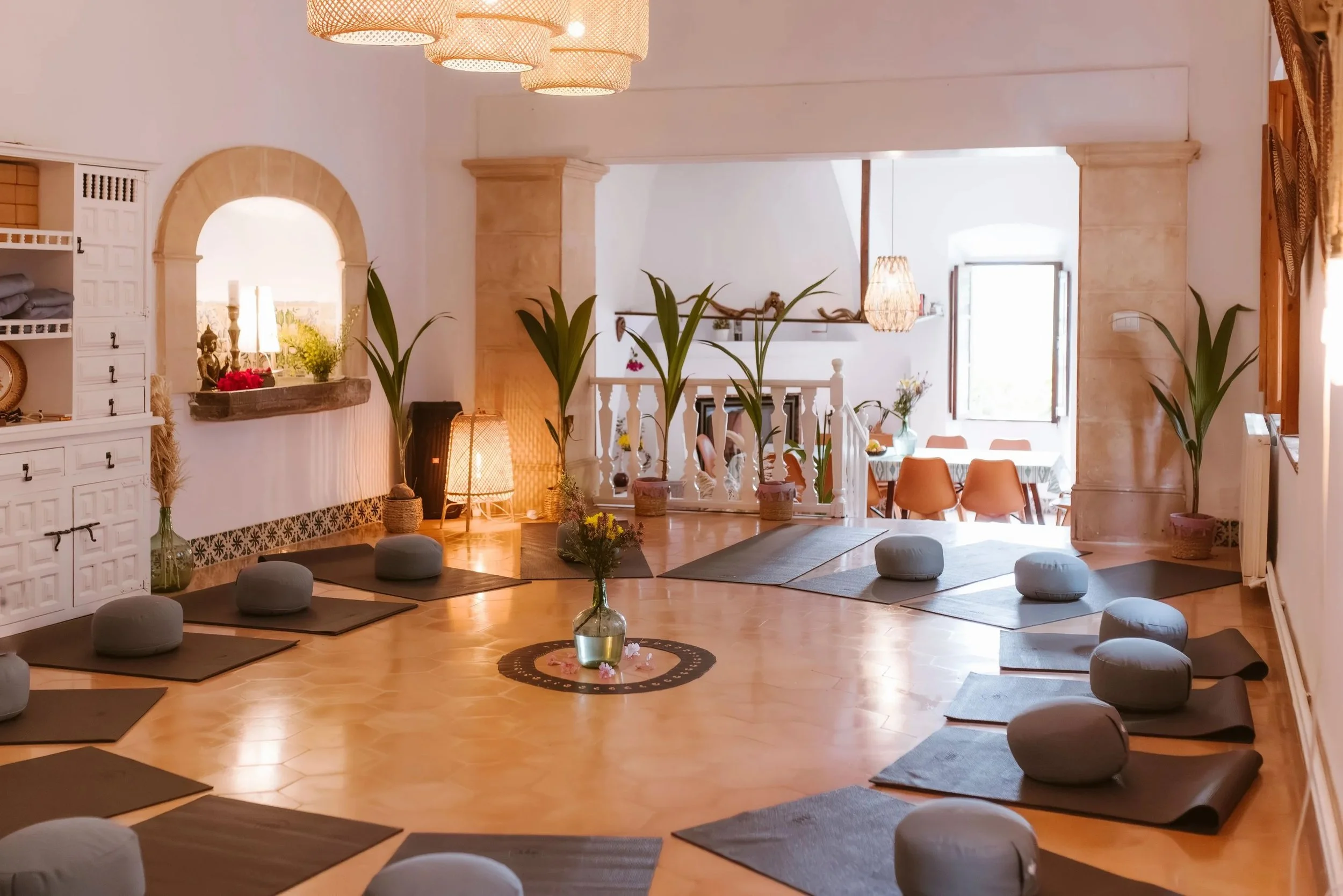 A spacious, well-lit room set up for yoga or meditation with mats, round pillows, and large green plants. There are decorative lamps and natural light coming through the windows, creating a peaceful atmosphere.