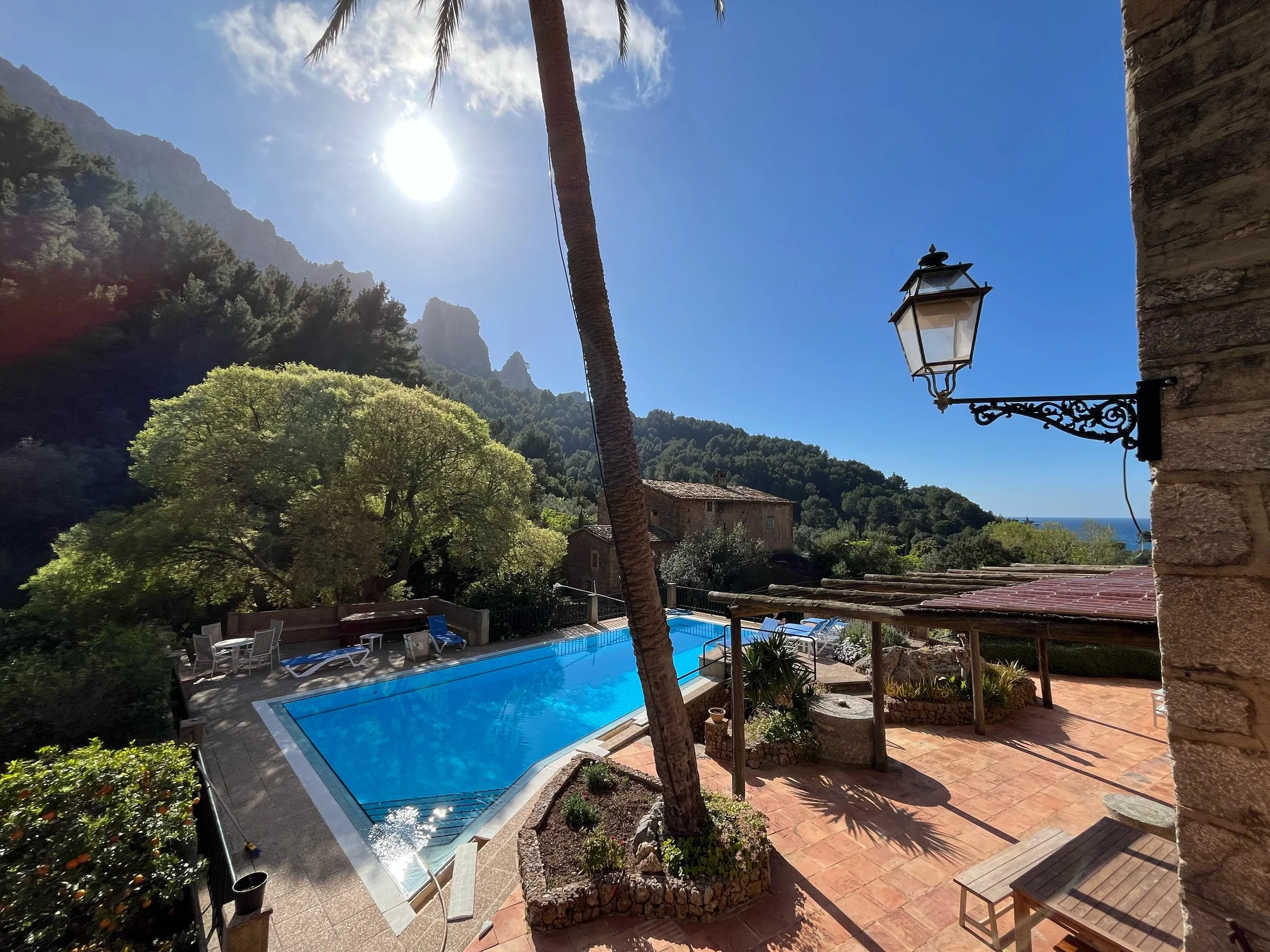 Sunny outdoor pool area with lounge chairs, surrounded by trees, mountains, and a clear blue sky, with a lamp post attached to a stone wall in the foreground.