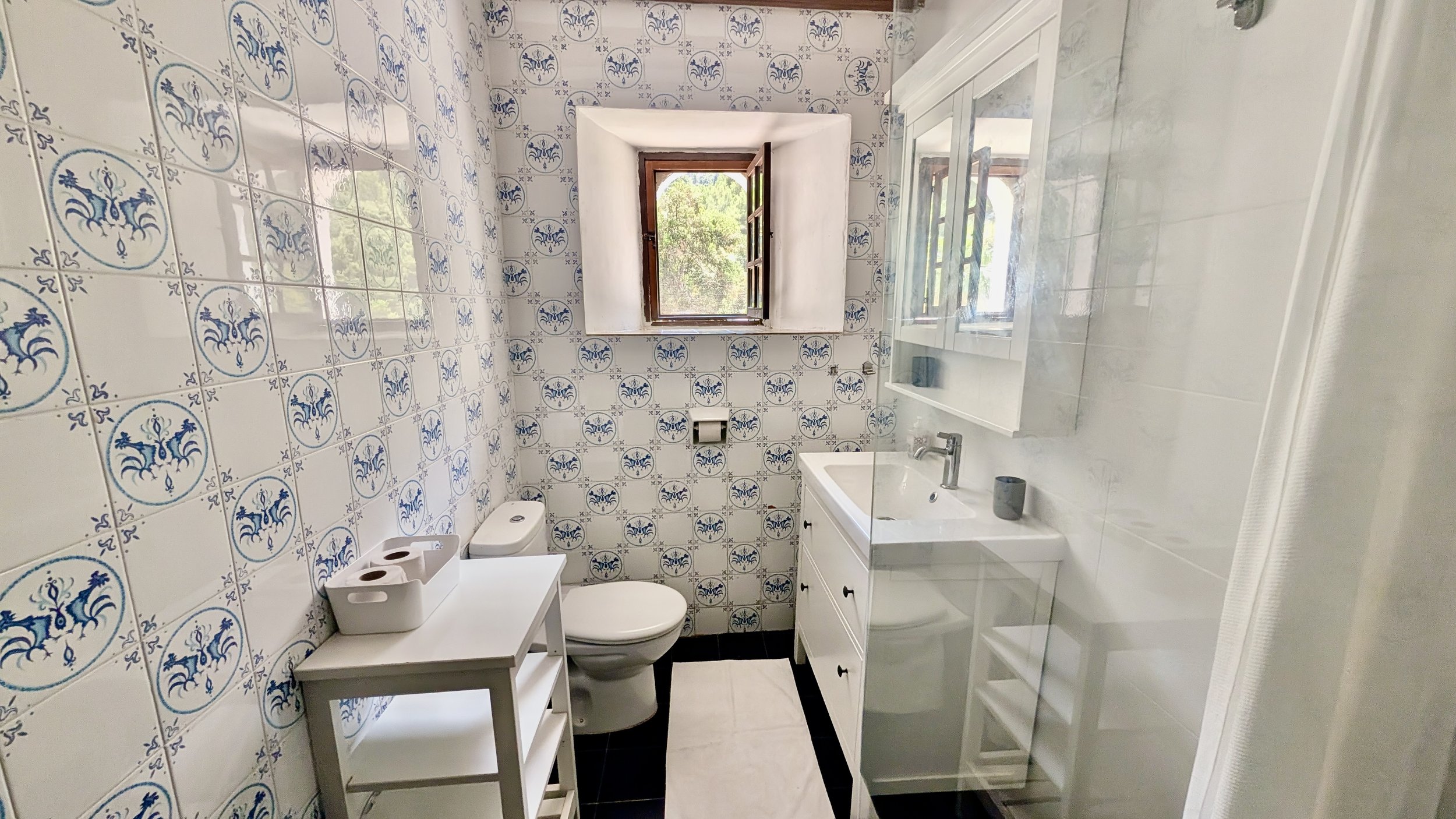 Small bathroom with patterned blue and white tile walls, a small window, toilet, white vanity with sink, mirror, and shelves.