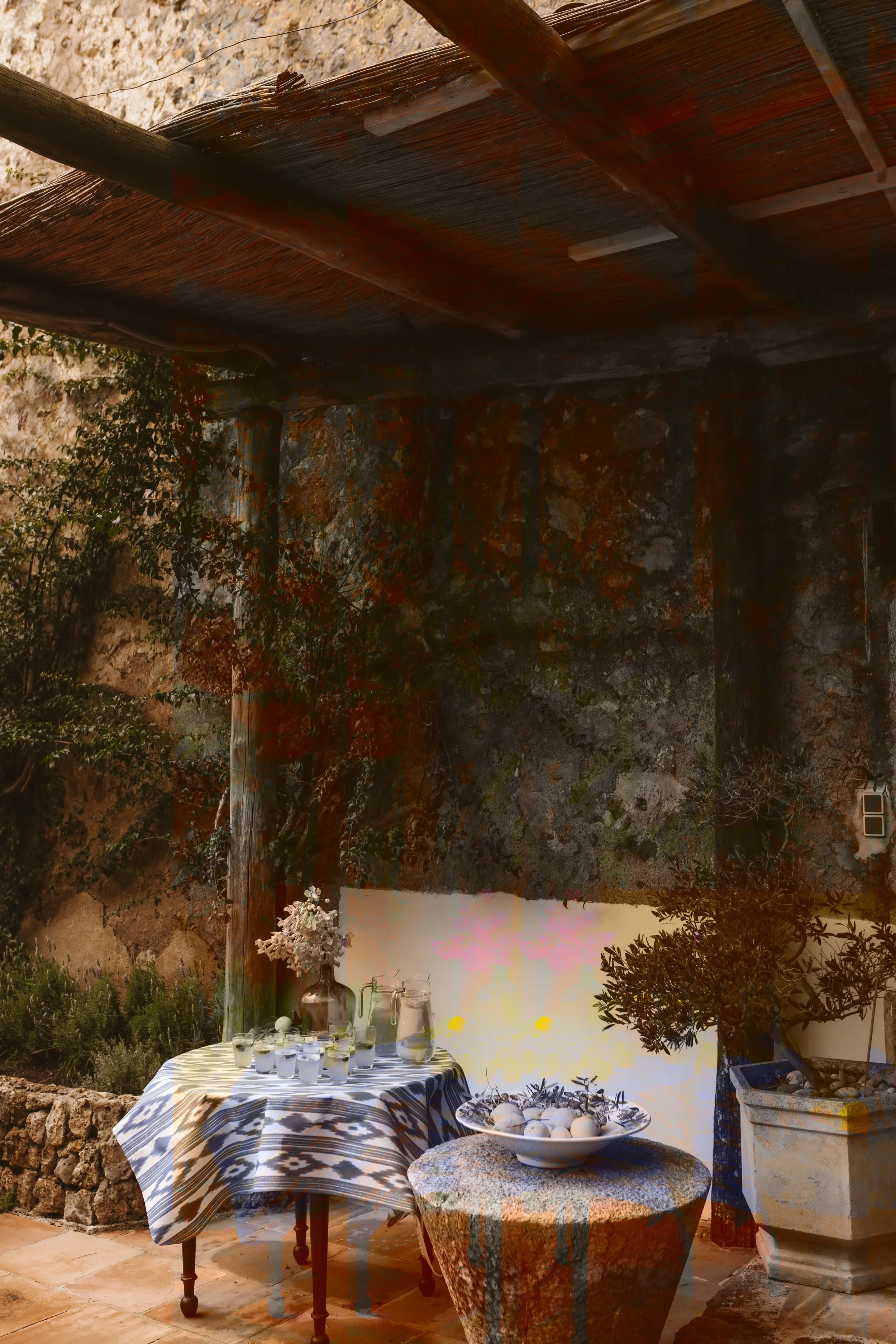 Outdoor patio with a round table covered with a patterned cloth, holding a glass pitcher, glasses, and a vase with flowers. Next to it, a smaller stone table has a bowl with white eggs and decorative branches. There is a potted plant in a large container and a textured stone wall in the background, with shaded sunlight.