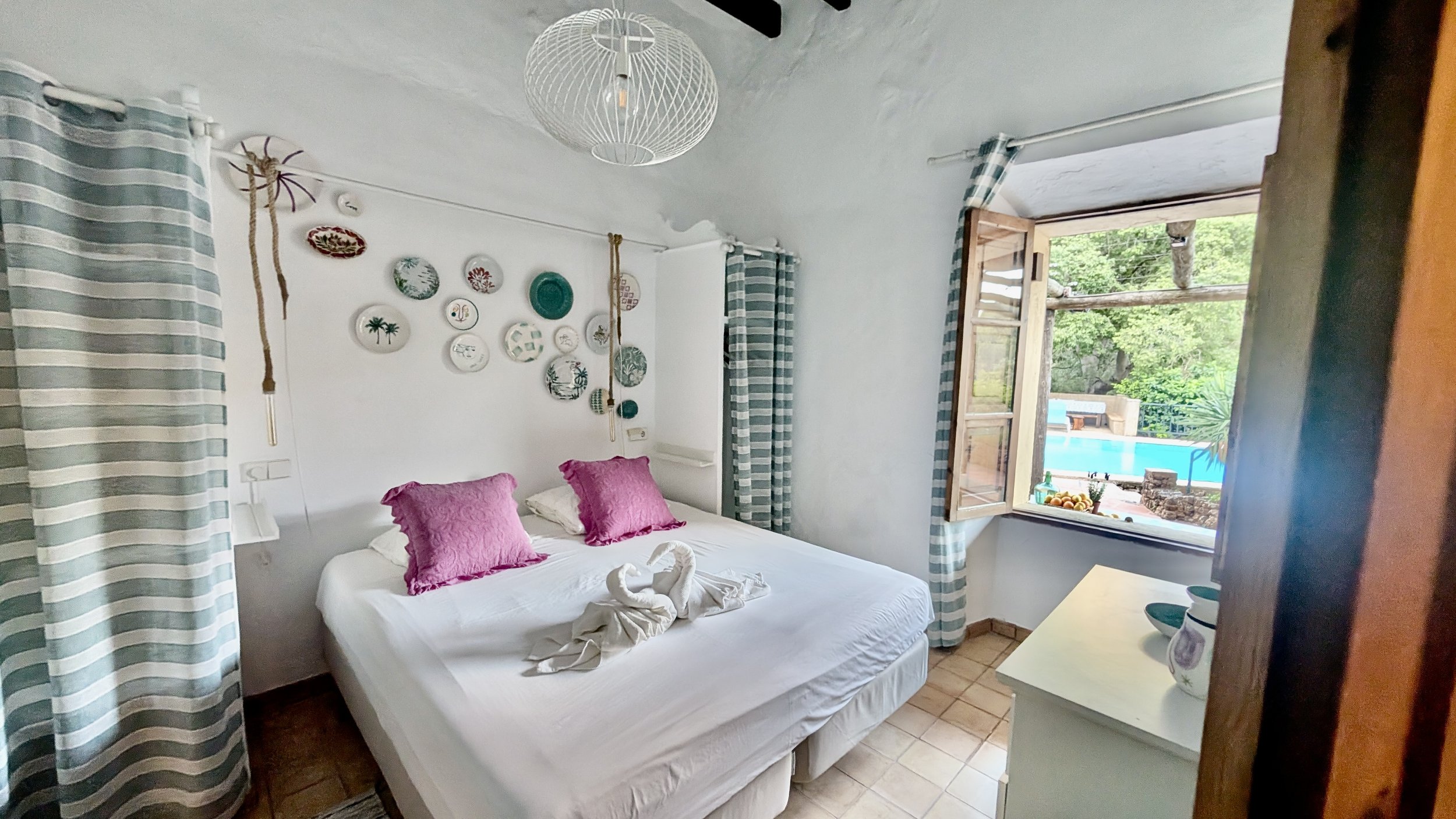 Cozy bedroom with white walls, decorated with a wall hanging of decorative plates, two pink pillows on a white bed, two towel swan sculptures, and a view of a pool outside the window surrounded by greenery.