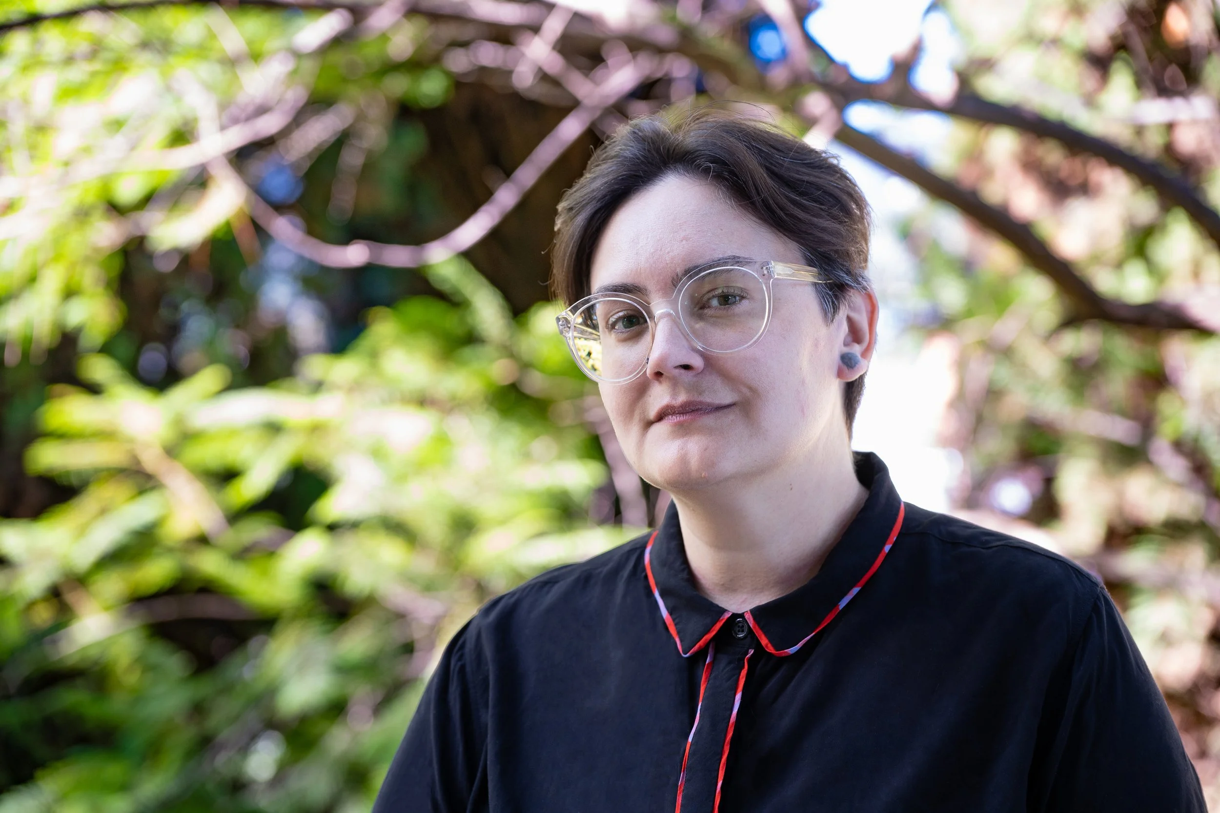 A medium close-up portrait of a person with short brown hair, wearing clear-framed glasses and a black collared shirt with red trim, smiling gently against a softly blurred background of green foliage.