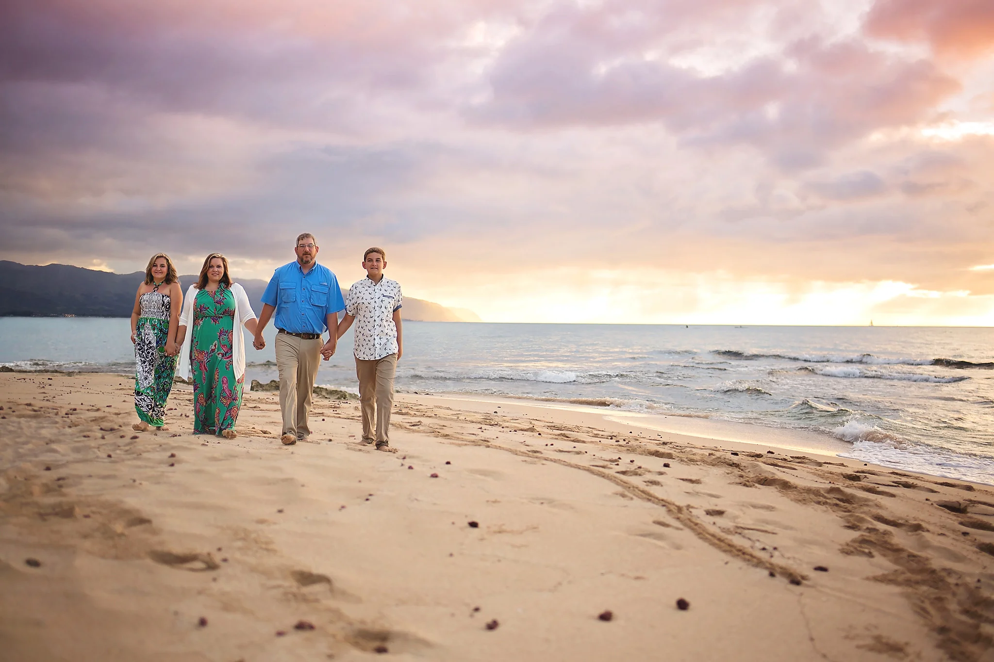 Family of 4 holding hands looking at the camera as they walk in a line along the beach at sunset. 