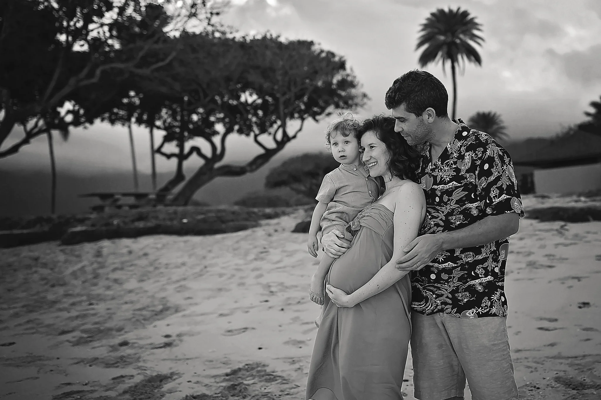 Black and white photo of a family of 3 standing on a beach looking out off into the distance. Kaneohe, Oahu, Hawaii.