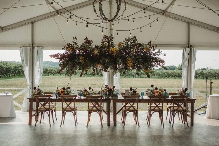 Two harvest tables set with plates and flowers with a large floral installation hanging above them