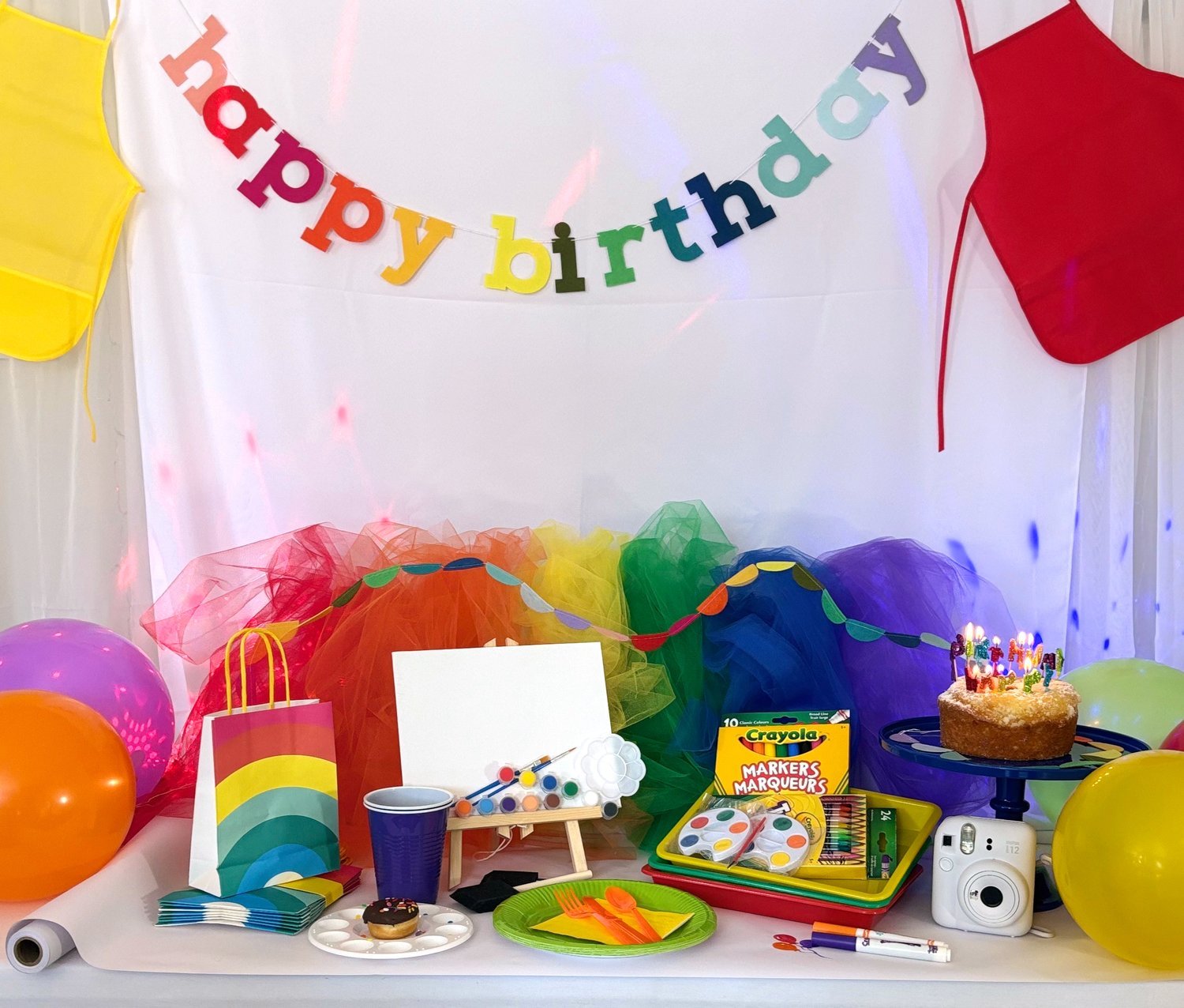Rainbow "Happy Birthday" banner above a table with art supplies, colourful ribbons, and balloons