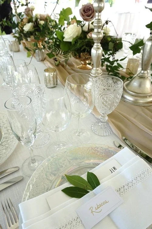 Place setting with four different glasses for drinks next gold table runner with greenery in vases