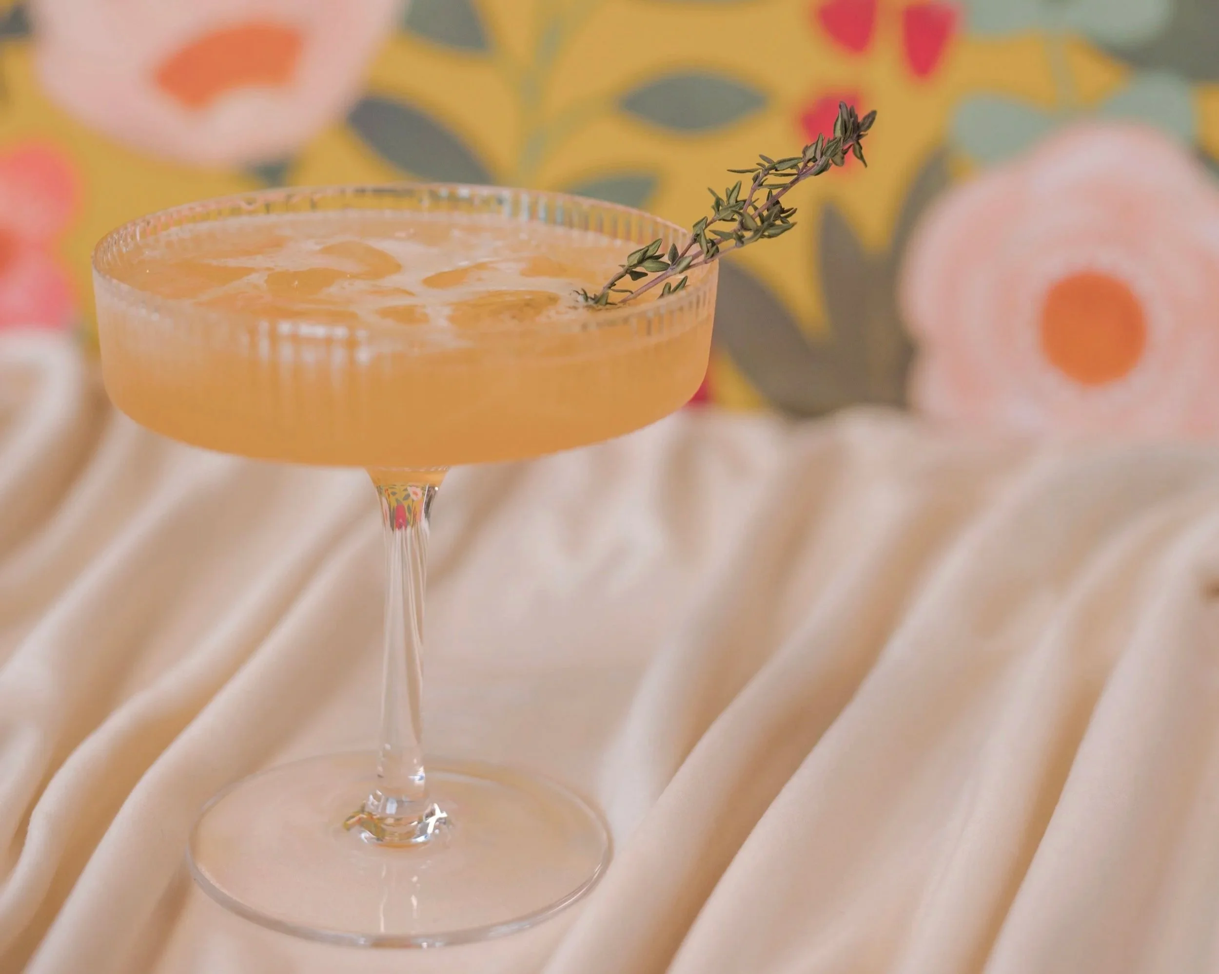 Cocktail with fresh thyme in a coupe glass against a green floral backdrop
