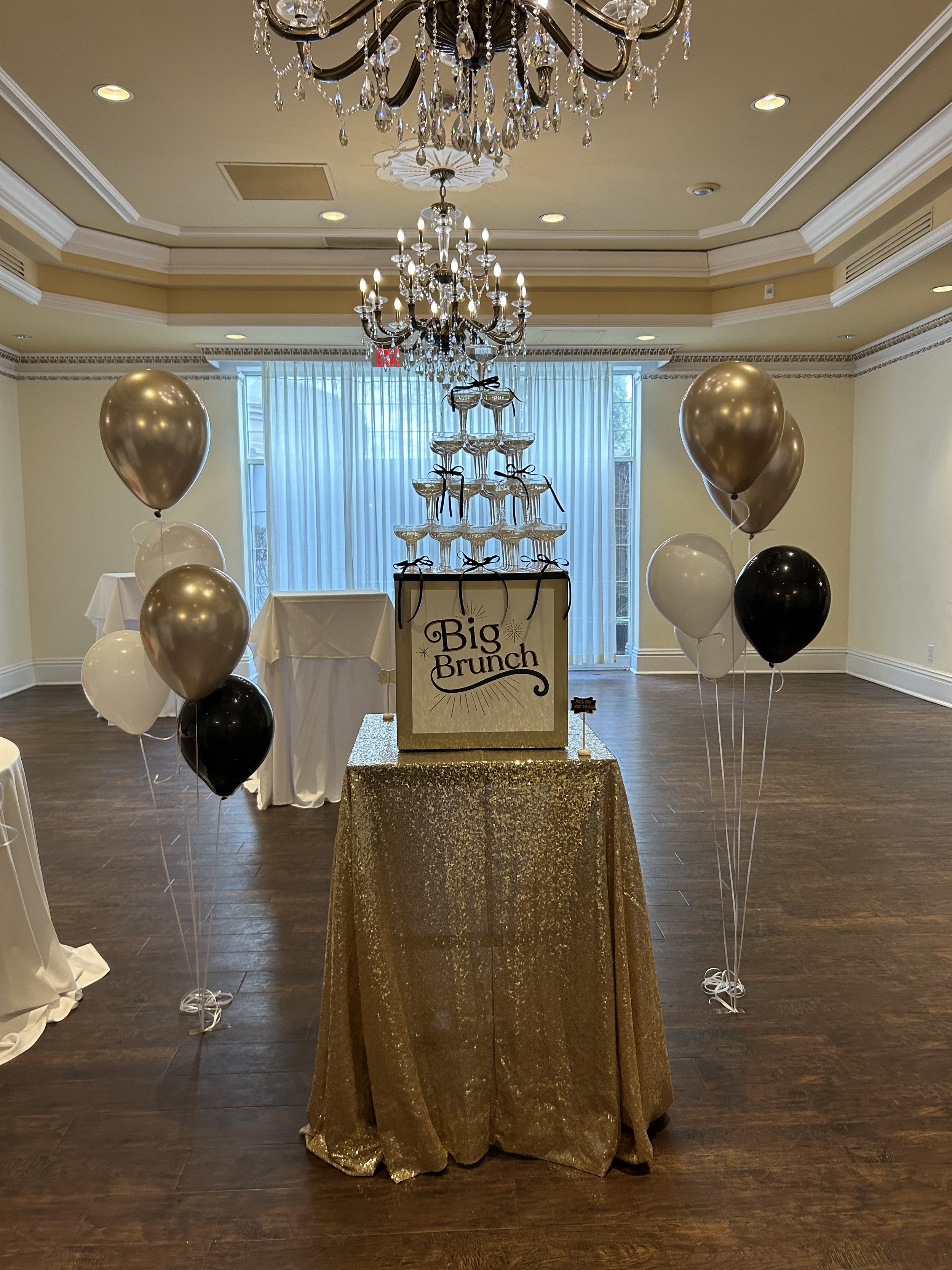 Champagne tower on a box that says "Big Brunch" with a sparkly gold linen with black, white, and gold balloons around it