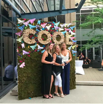 Three people standing in front of moss photo backdrop is "COOL" spelled out in corks with paper butterfly scattered across moss
