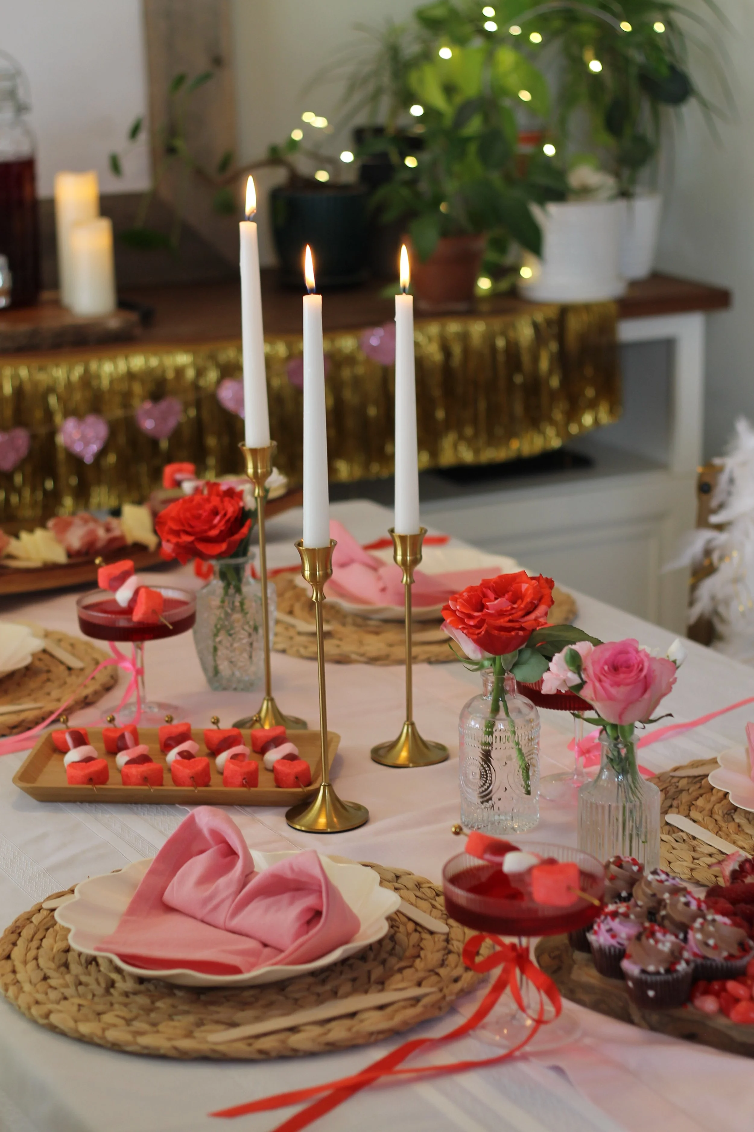 Three candlesticks burning on table with desserts, roses, and place settings with pink napkins folded into hearts