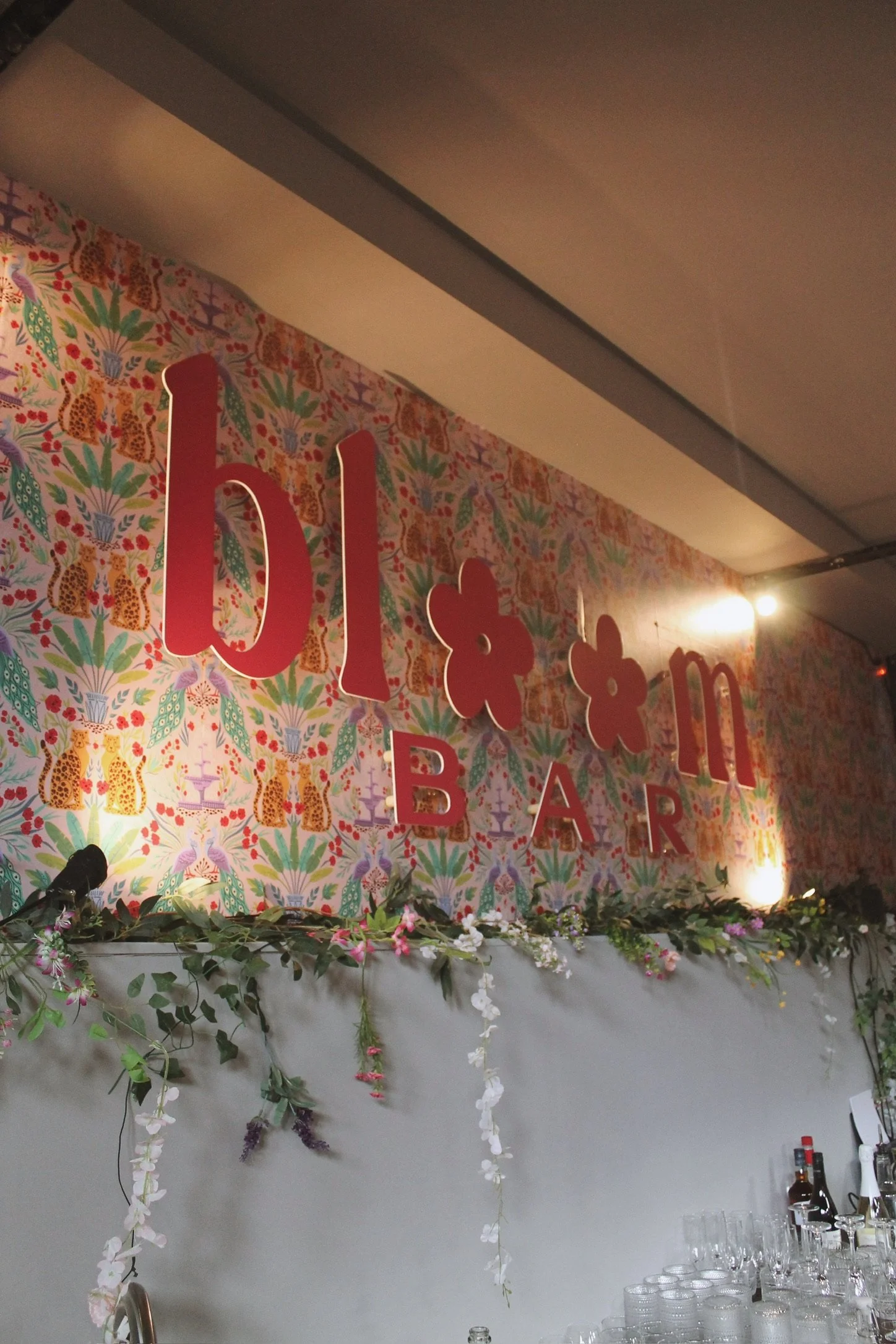 Pink jungle themed wallpaper with a 'bloom bar' sign being illuminated by spotlights and flower stands
